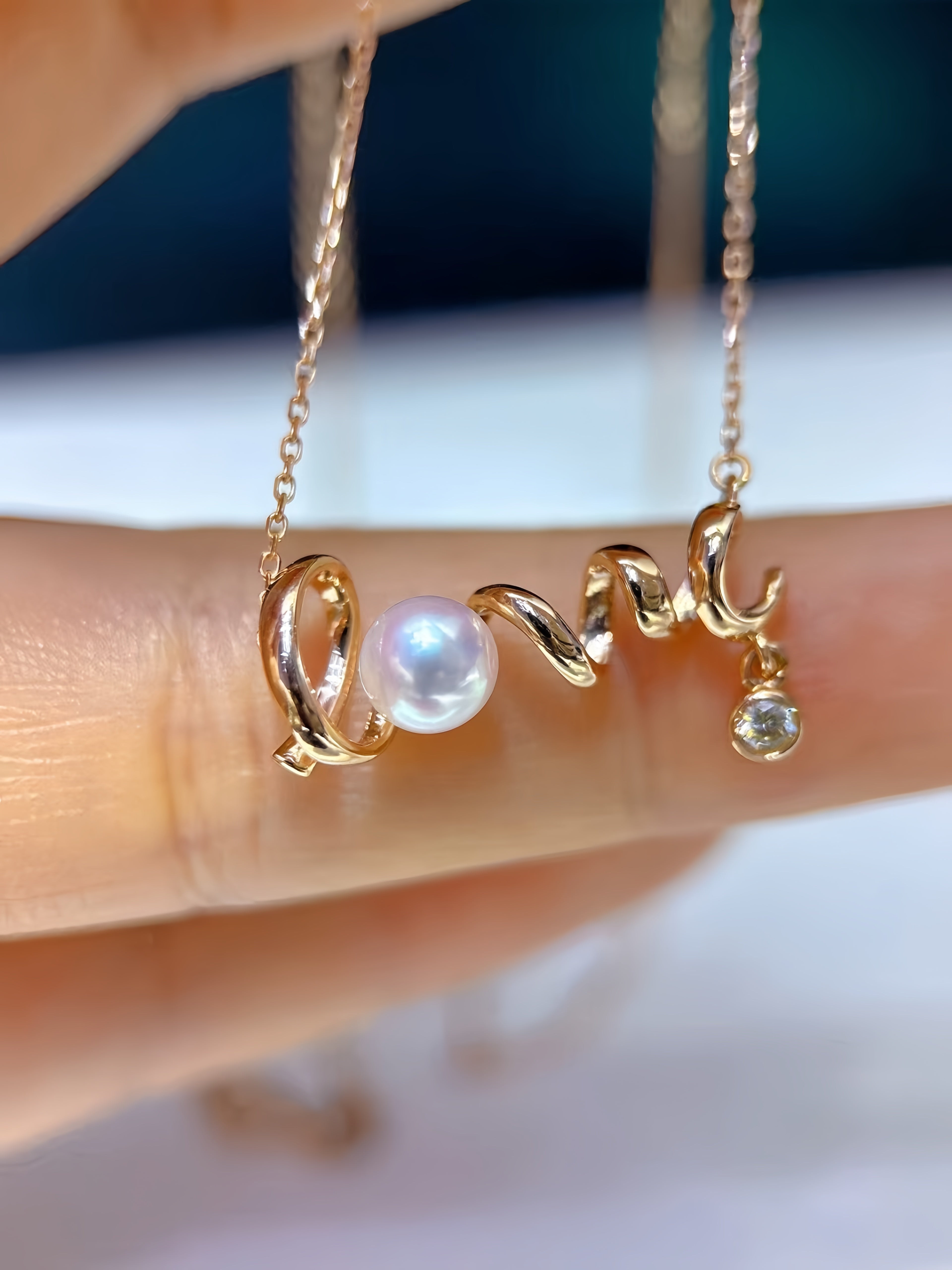 A close-up of a delicate gold necklace with a curved, twisted design featuring a white pearl and a small clear gemstone, held between fingers.