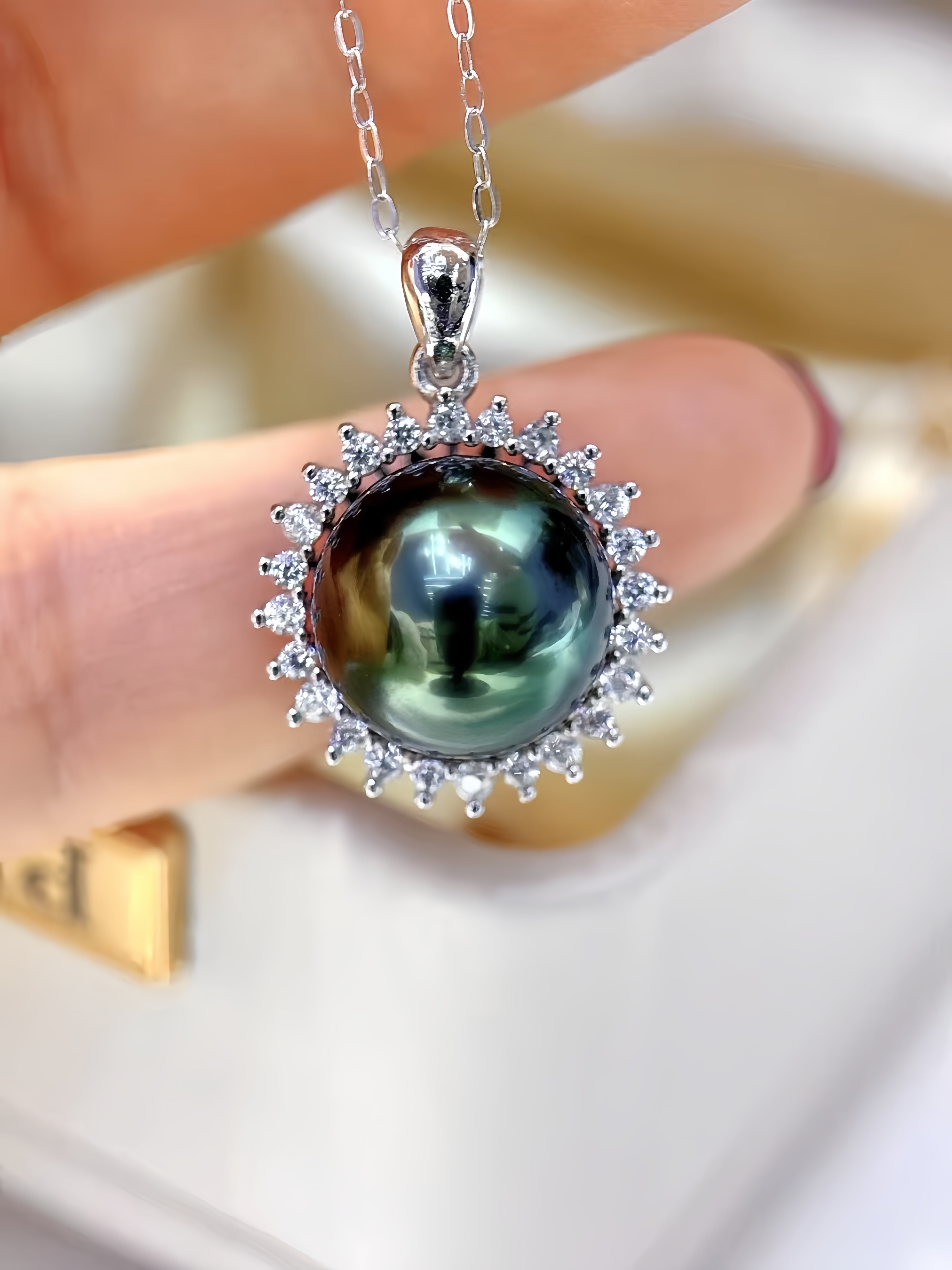 A close-up of a silver pendant necklace featuring a large iridescent green pearl surrounded by small clear stones, held between fingers.