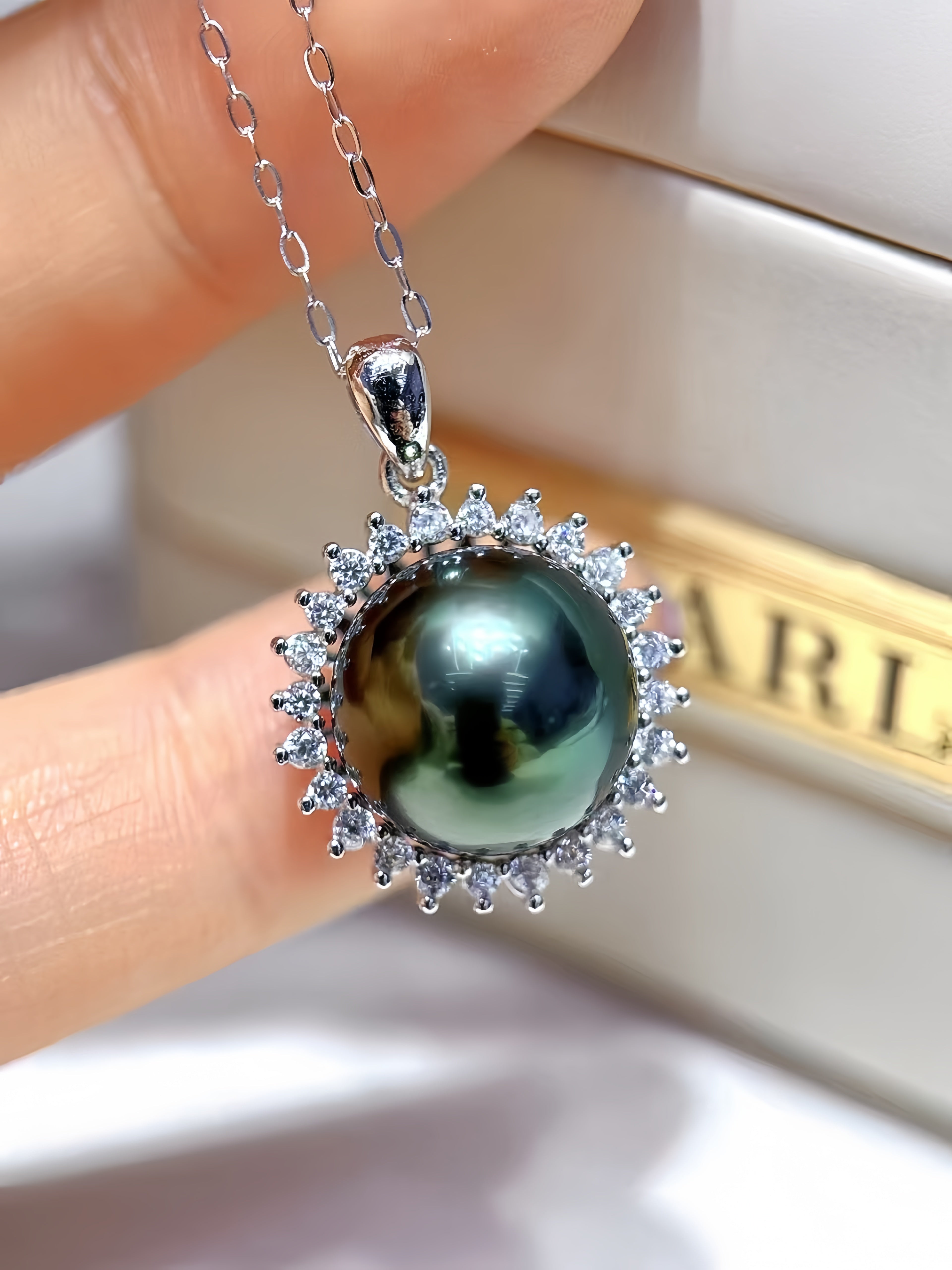A close-up of a silver pendant necklace held between fingers, featuring a round dark green-black pearl surrounded by small clear stones, with a blurred jewelry box in the background.