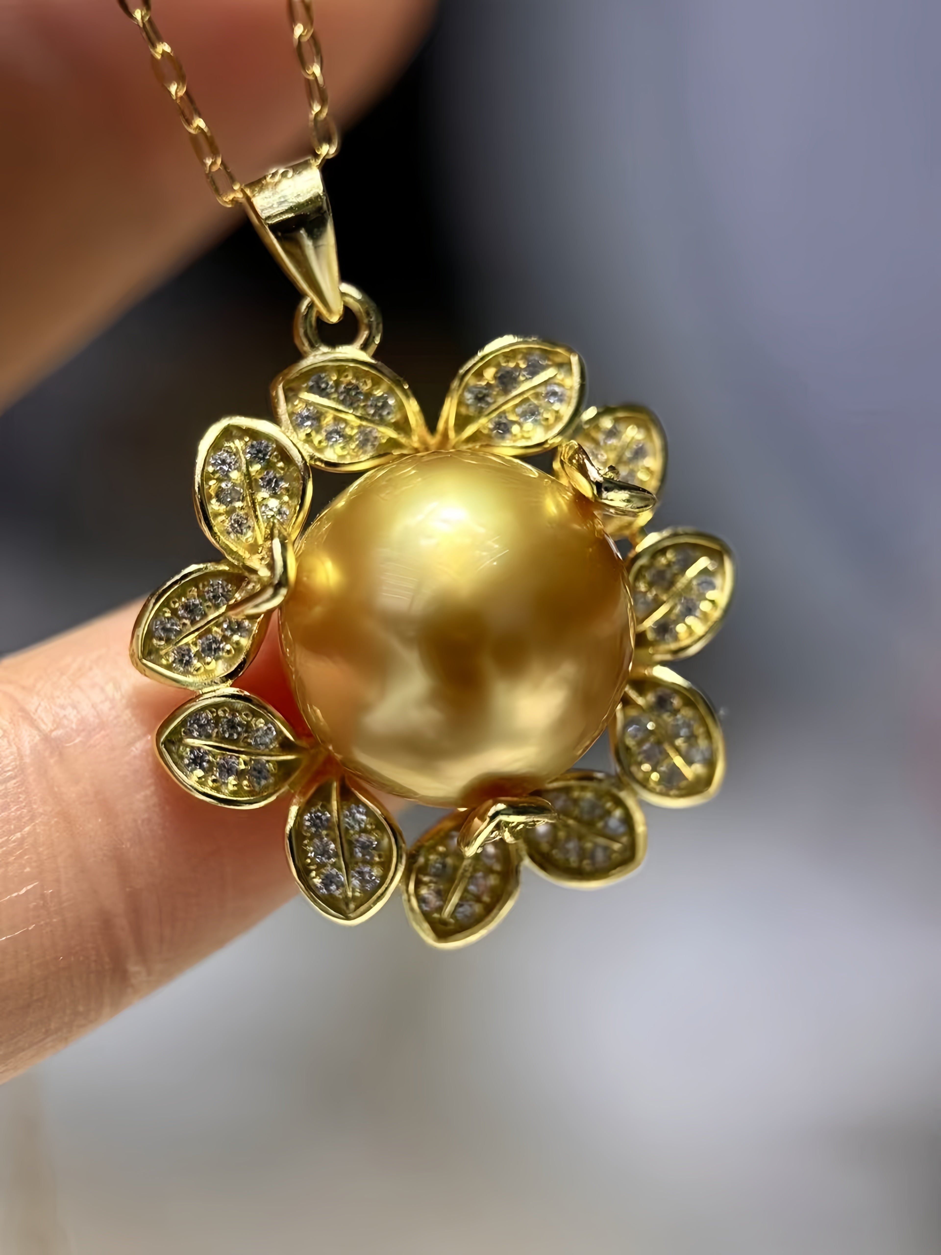 A gold pendant necklace featuring a large round gold pearl in the center, surrounded by a flower-like halo of small gold leaves paved with clear stones, held between fingers.