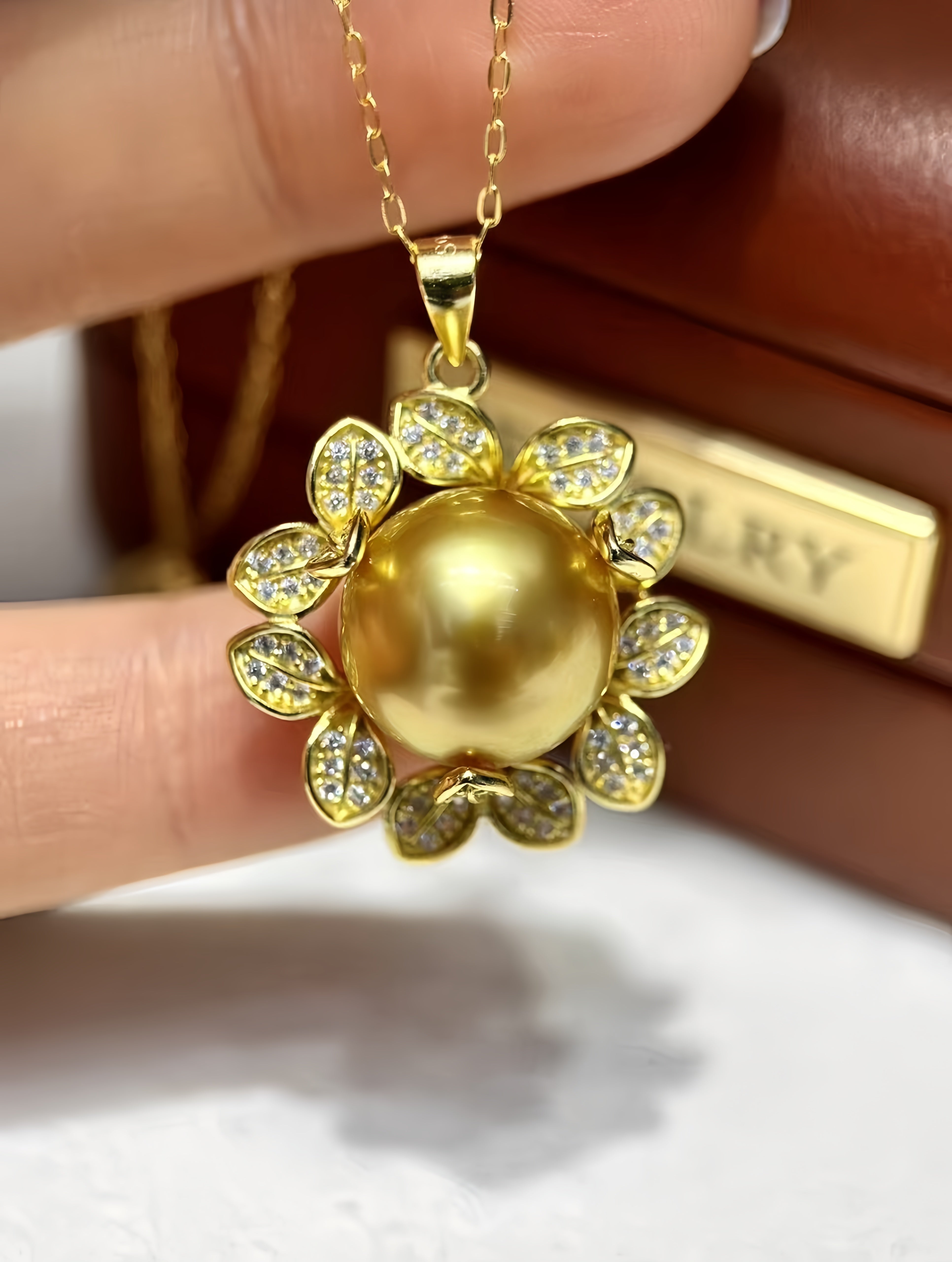 A gold pendant necklace featuring a large round pearl-like center surrounded by leaf-shaped settings set with small clear stones, held between fingers against a brown jewelry box background.