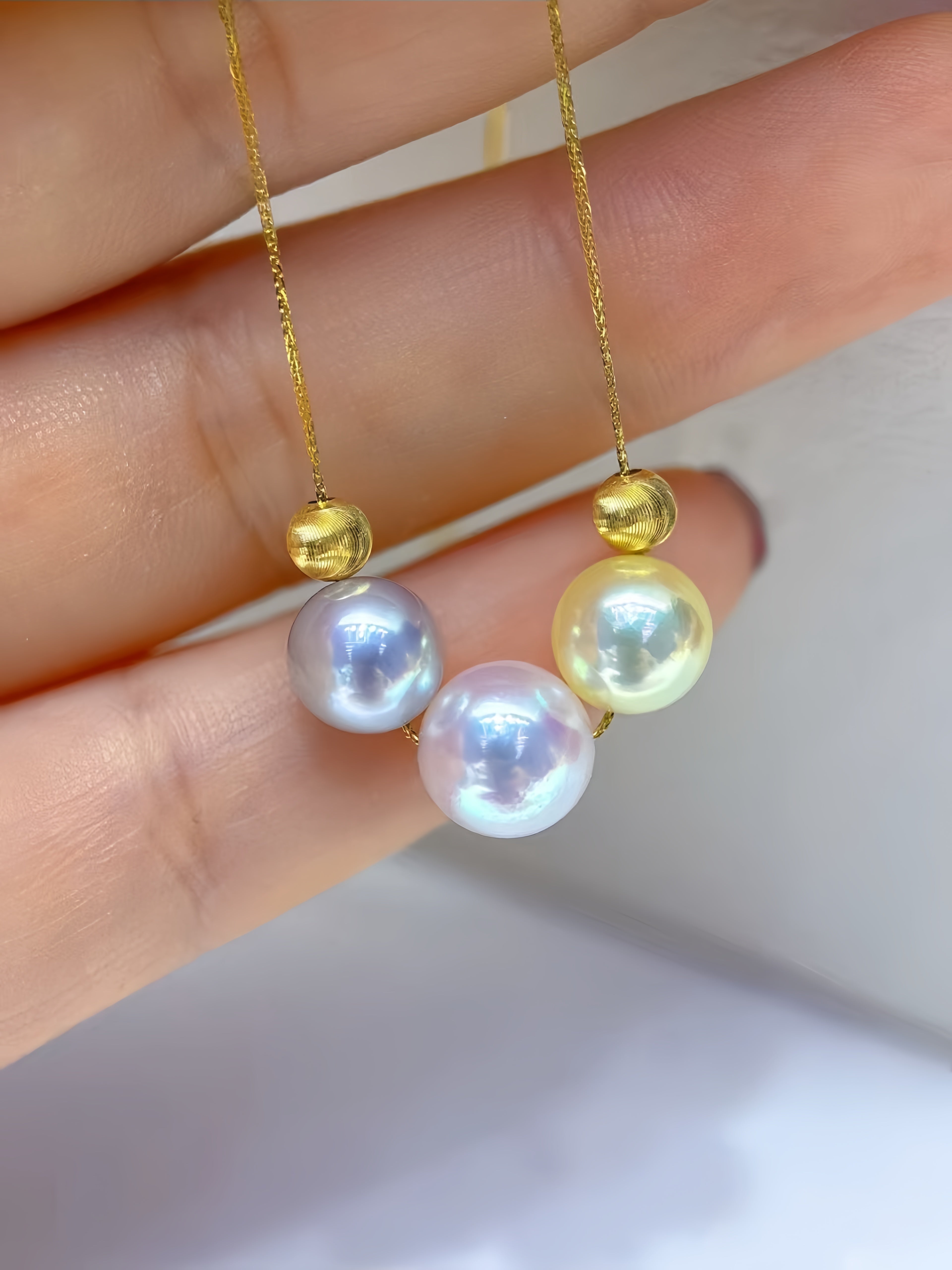 A close-up of a hand holding a delicate gold necklace with three iridescent freshwater pearls of different colors: white, pale purple, and pale yellow, each accented by a small gold bead on a fine gold chain.