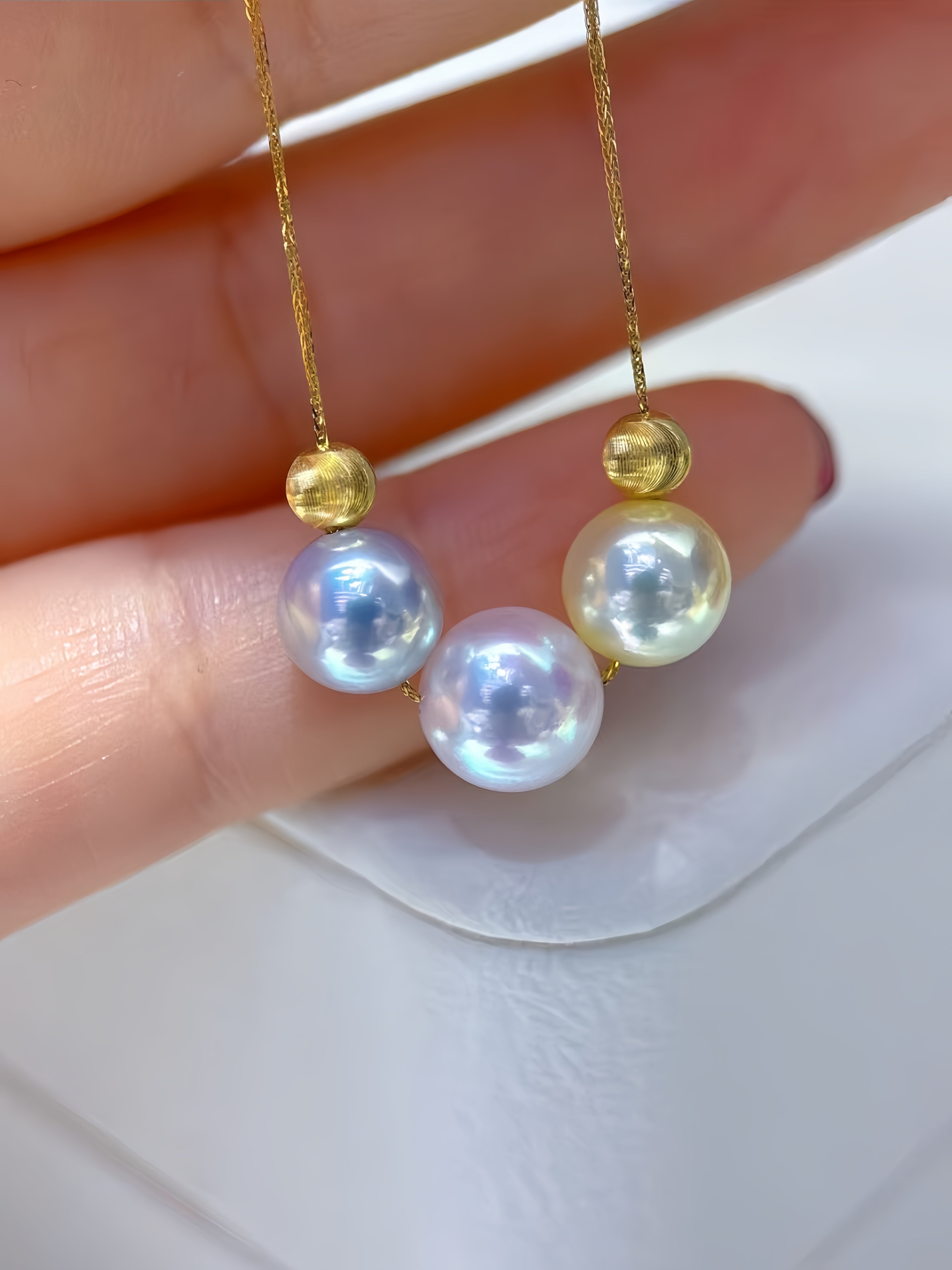 A close-up of a hand holding a pair of gold necklace pendants with round iridescent pearl-like beads and small gold spacer beads, against a soft white background.