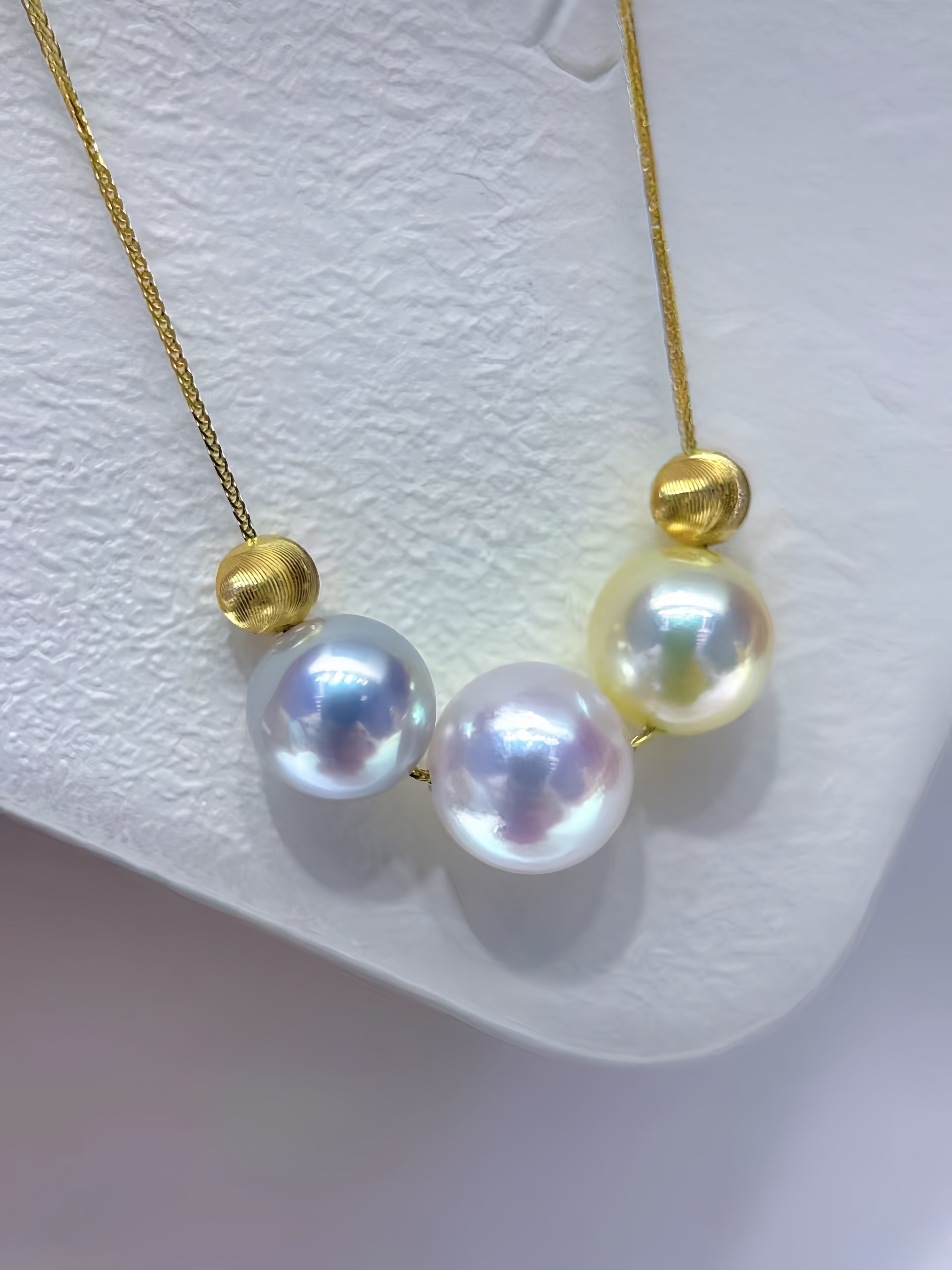 A gold chain necklace featuring three large, iridescent freshwater pearls in white, pale yellow, and light blue, accented by small gold beads, displayed on a textured white surface.