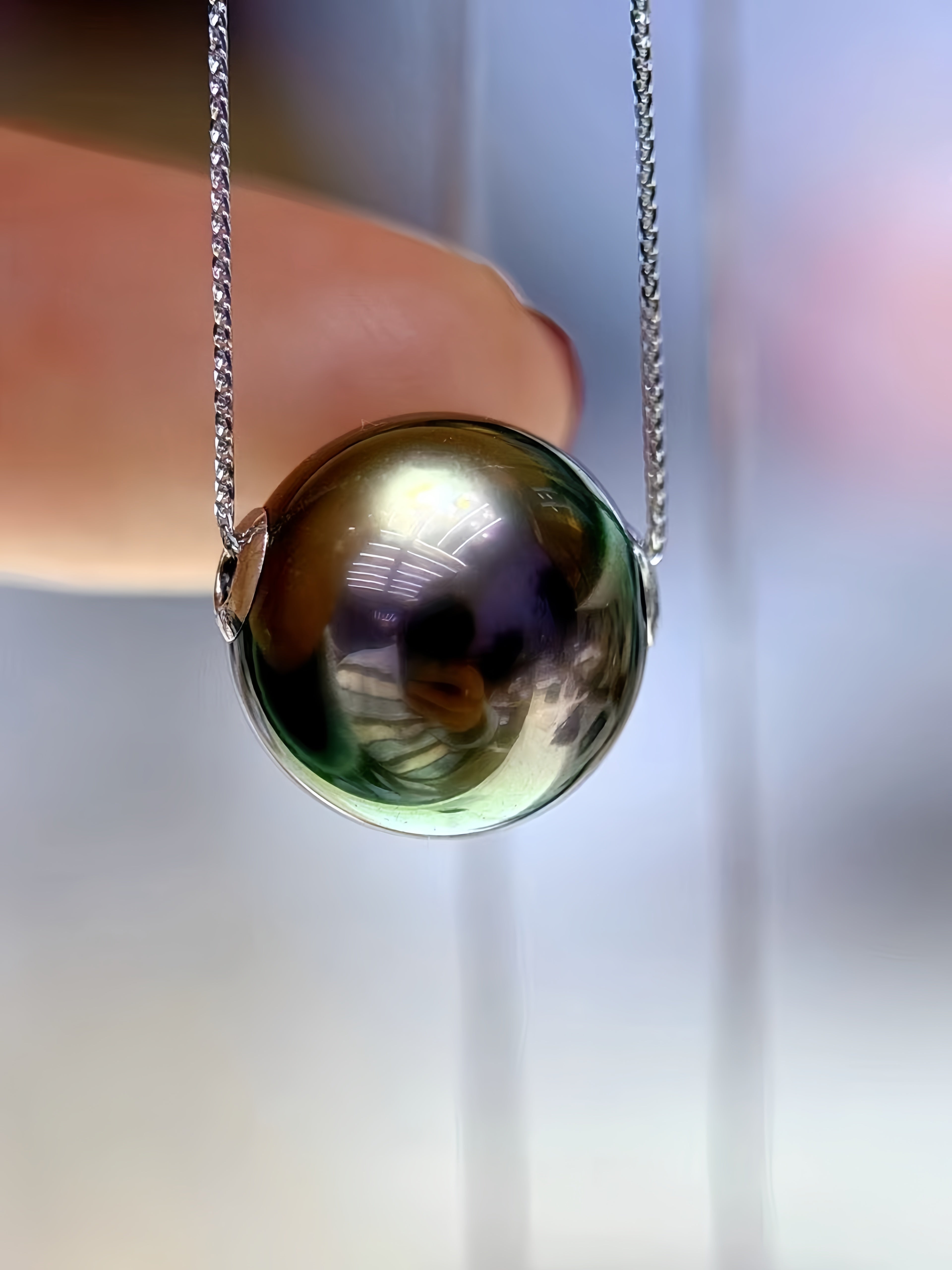 A close-up of a round iridescent pearl pendant on a silver chain, held by a person's finger against a softly blurred light background.