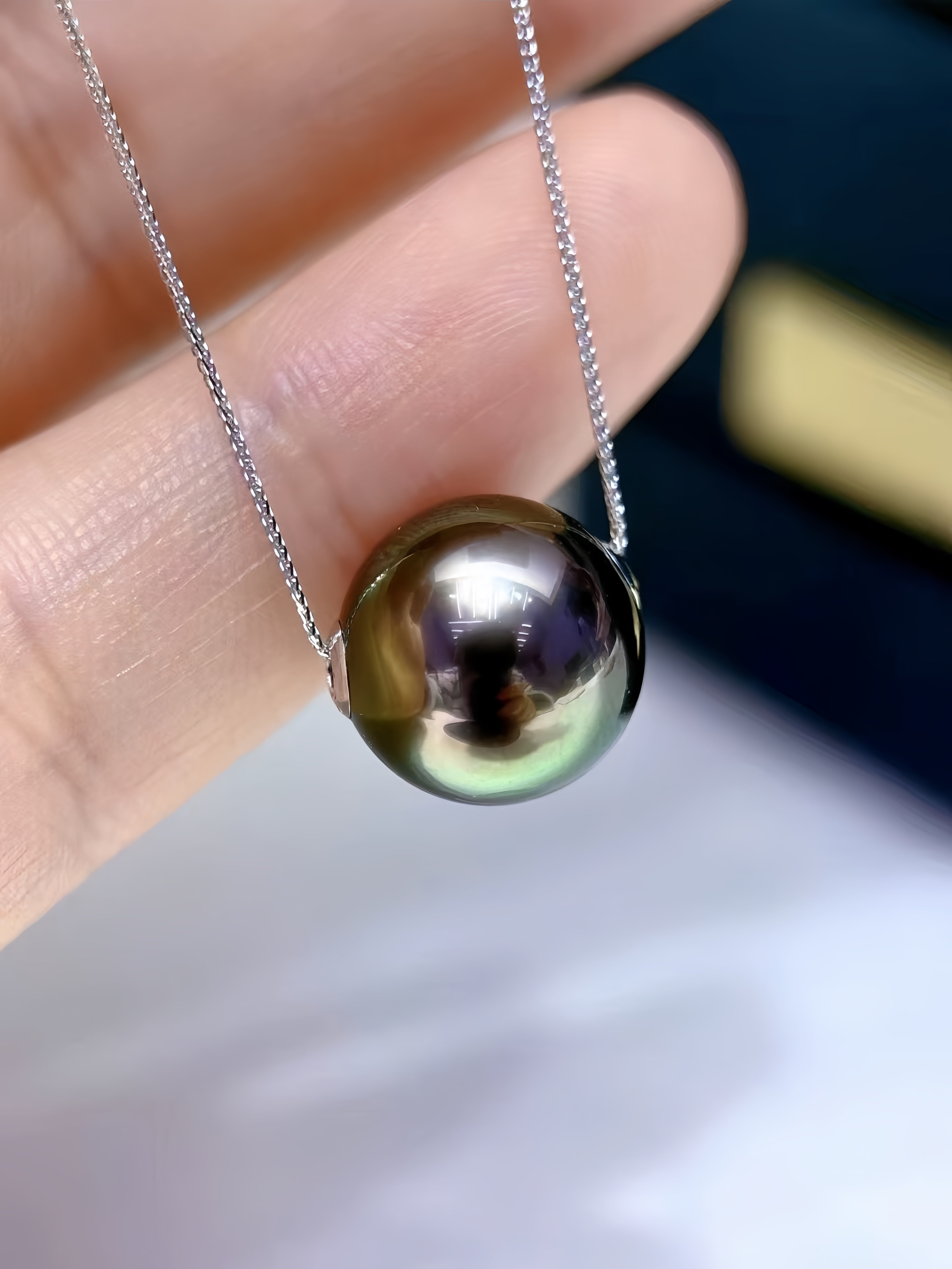 A close-up of a round iridescent pearl pendant on a delicate silver chain, held between fingers against a blurred dark background.