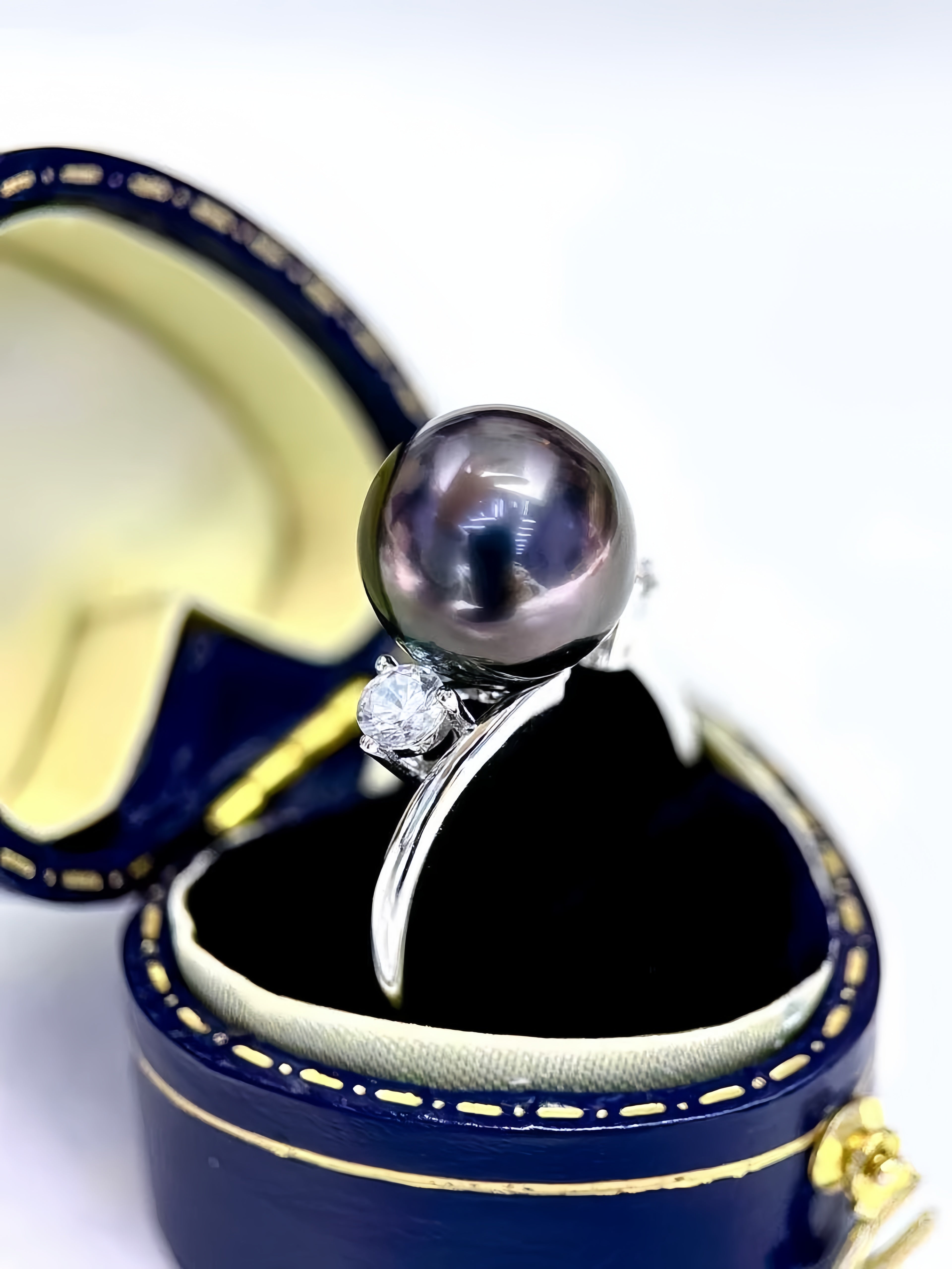 A dark gray freshwater pearl ring with a small clear diamond accent, displayed in an open navy blue jewelry box with gold trim.