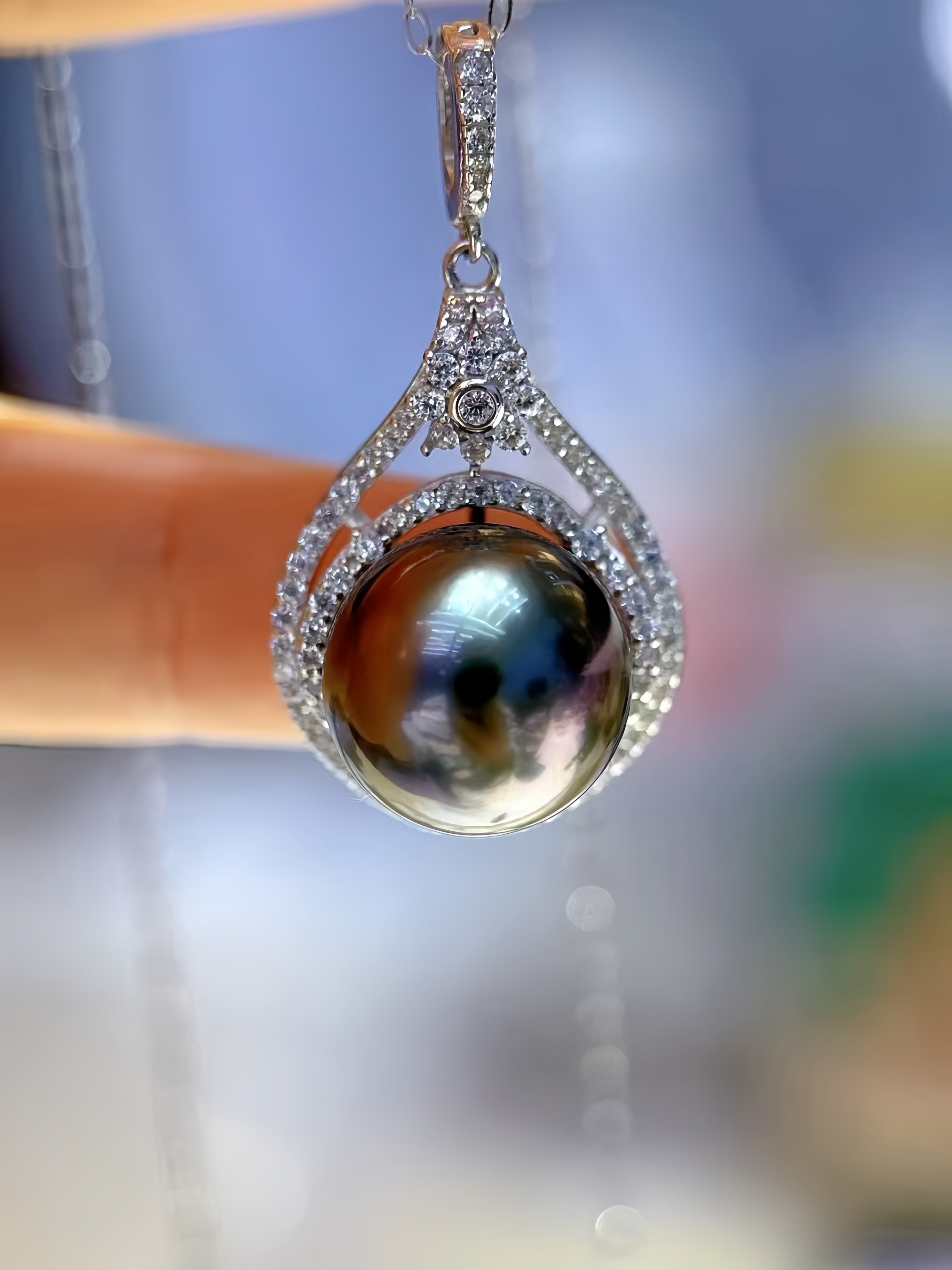 A close-up of a teardrop-shaped pendant featuring a large iridescent dark pearl surrounded by a silver-toned pavé setting, held by a finger against a blurred background.