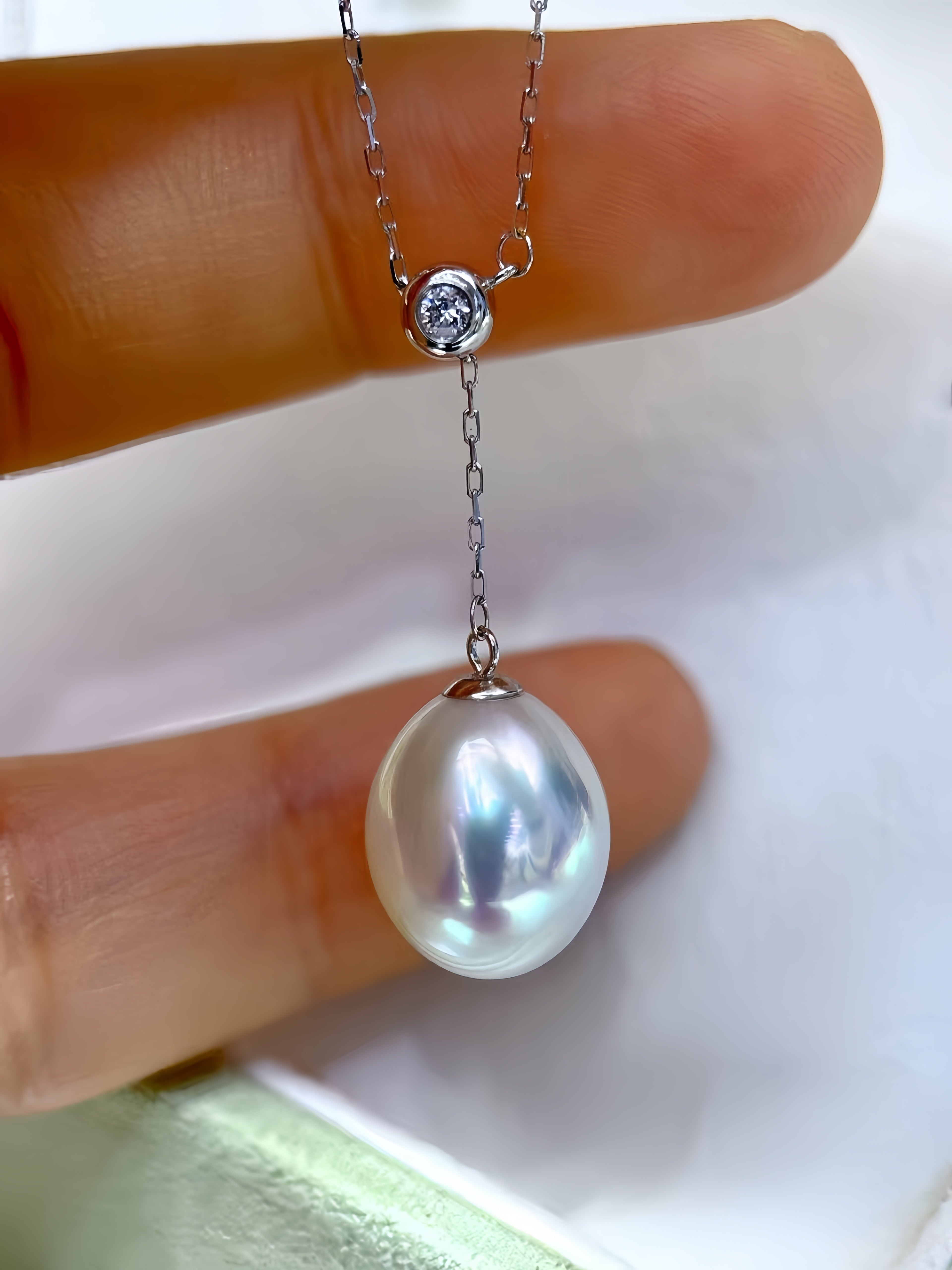 A close-up of a silver pendant necklace held between fingers. The necklace features a large, lustrous white pearl pendant hanging from a delicate silver chain, with a small round clear gemstone bezel-set above it.