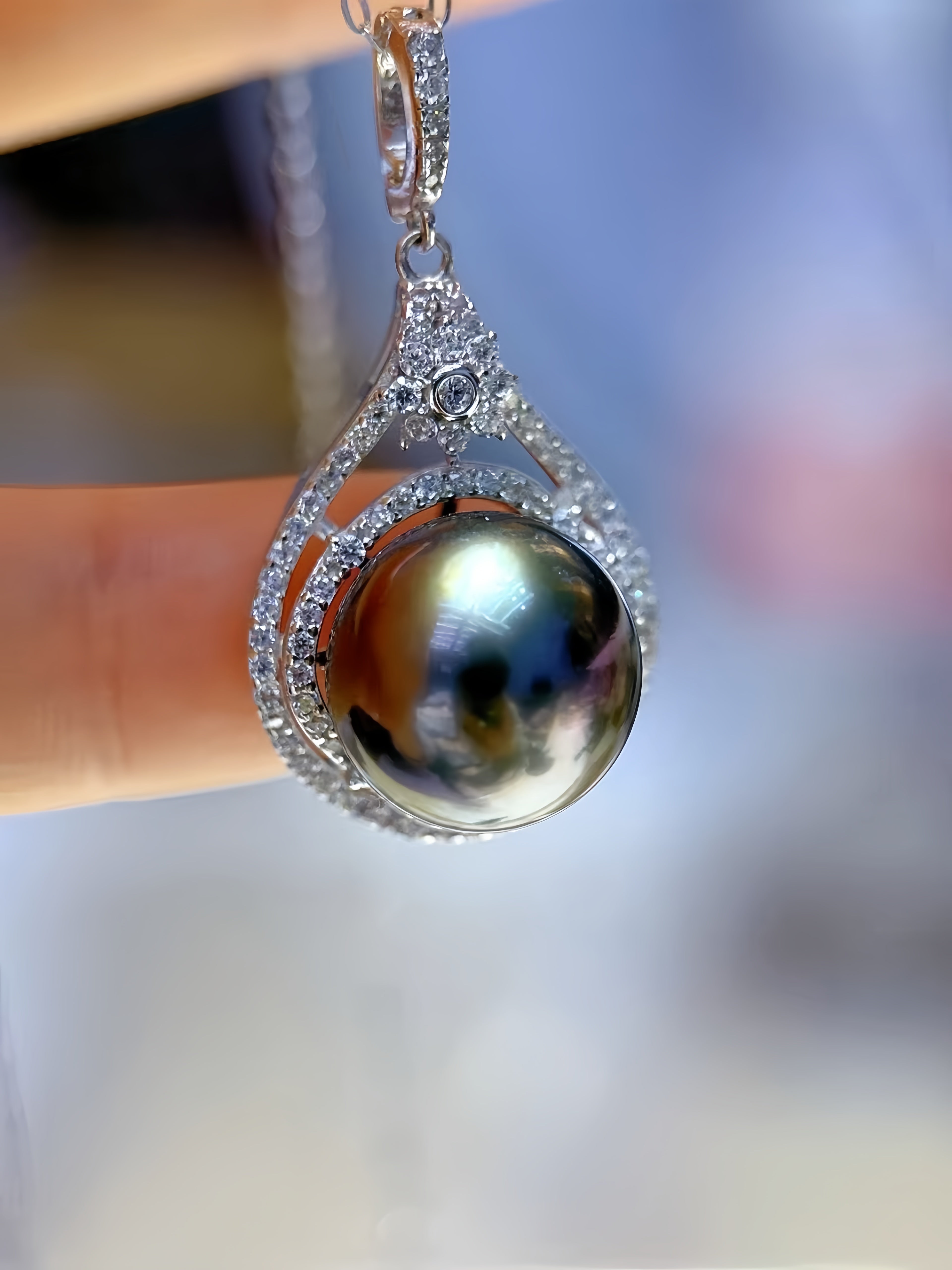 A close-up of a teardrop-shaped pendant necklace featuring a large iridescent dark pearl surrounded by a silver-toned halo of small clear stones, held against a blurred background.