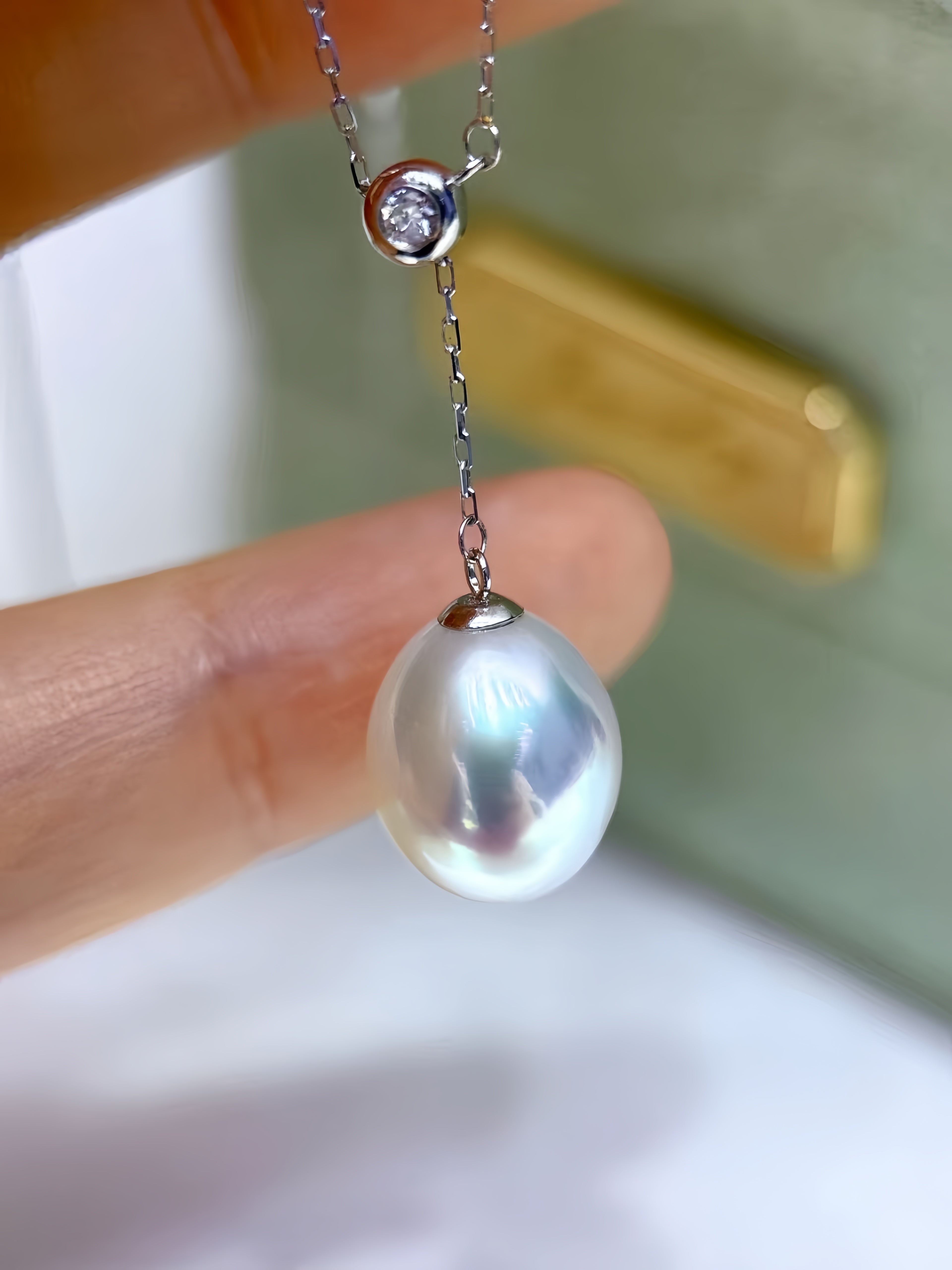A close-up of a hand holding a silver necklace with a large, lustrous white pearl pendant and a small clear gemstone accent.