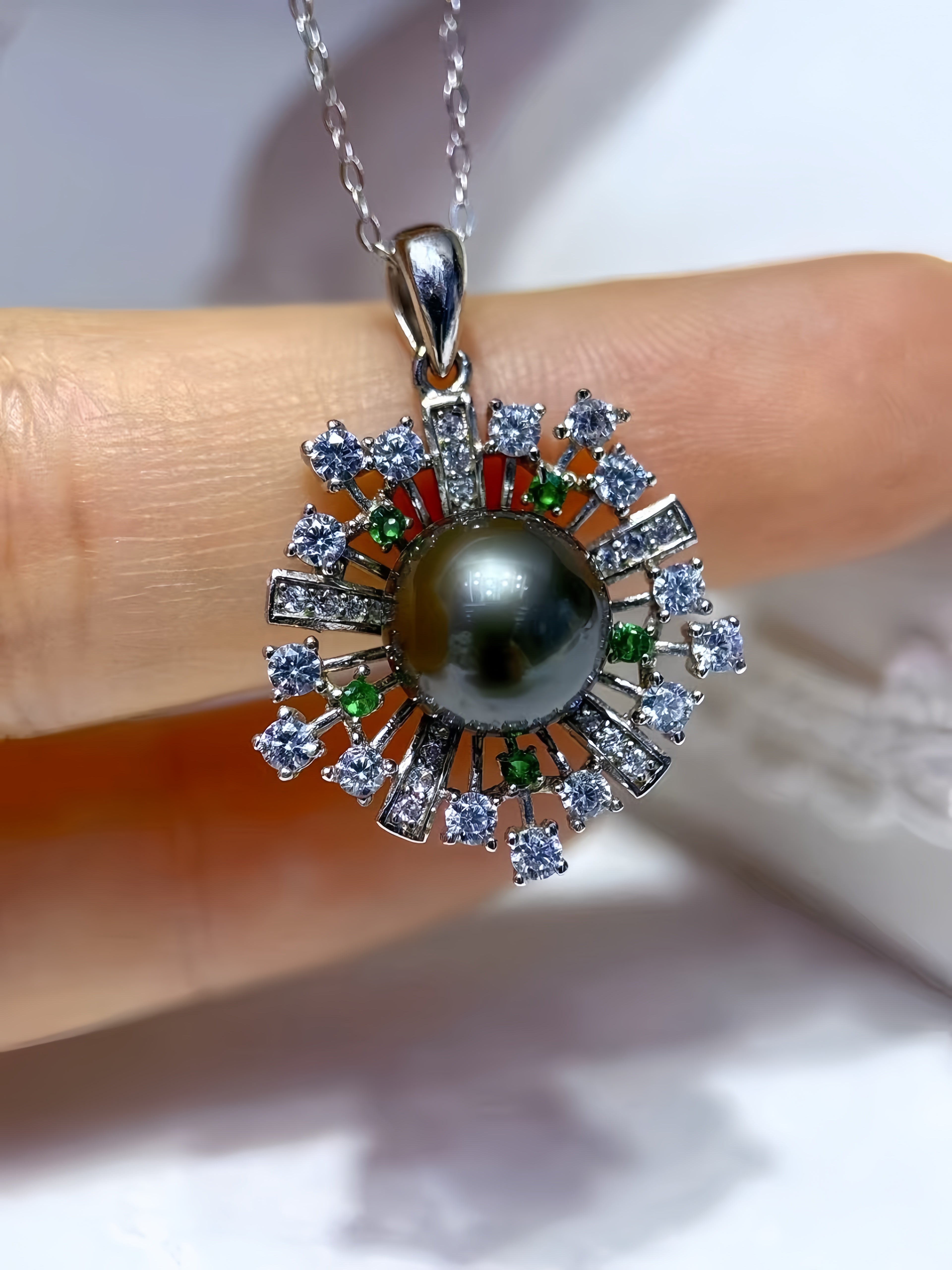 A close-up of a silver pendant necklace with a dark gray pearl center, surrounded by clear stones and small green and red accent stones, held against a finger.