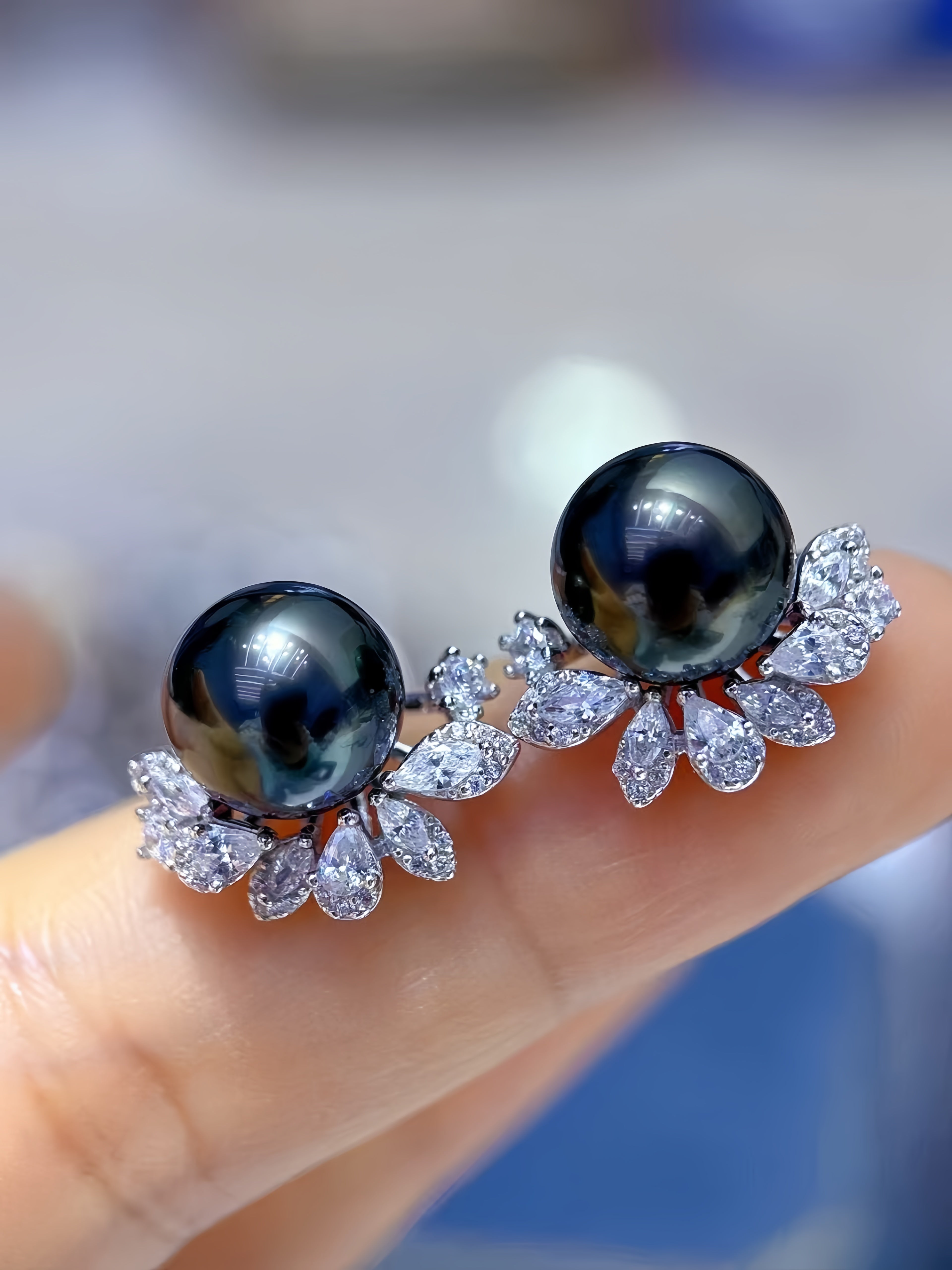 A pair of stud earrings with large round black pearls and silver-toned leaf clusters set with clear stones, shown resting on a person's finger.