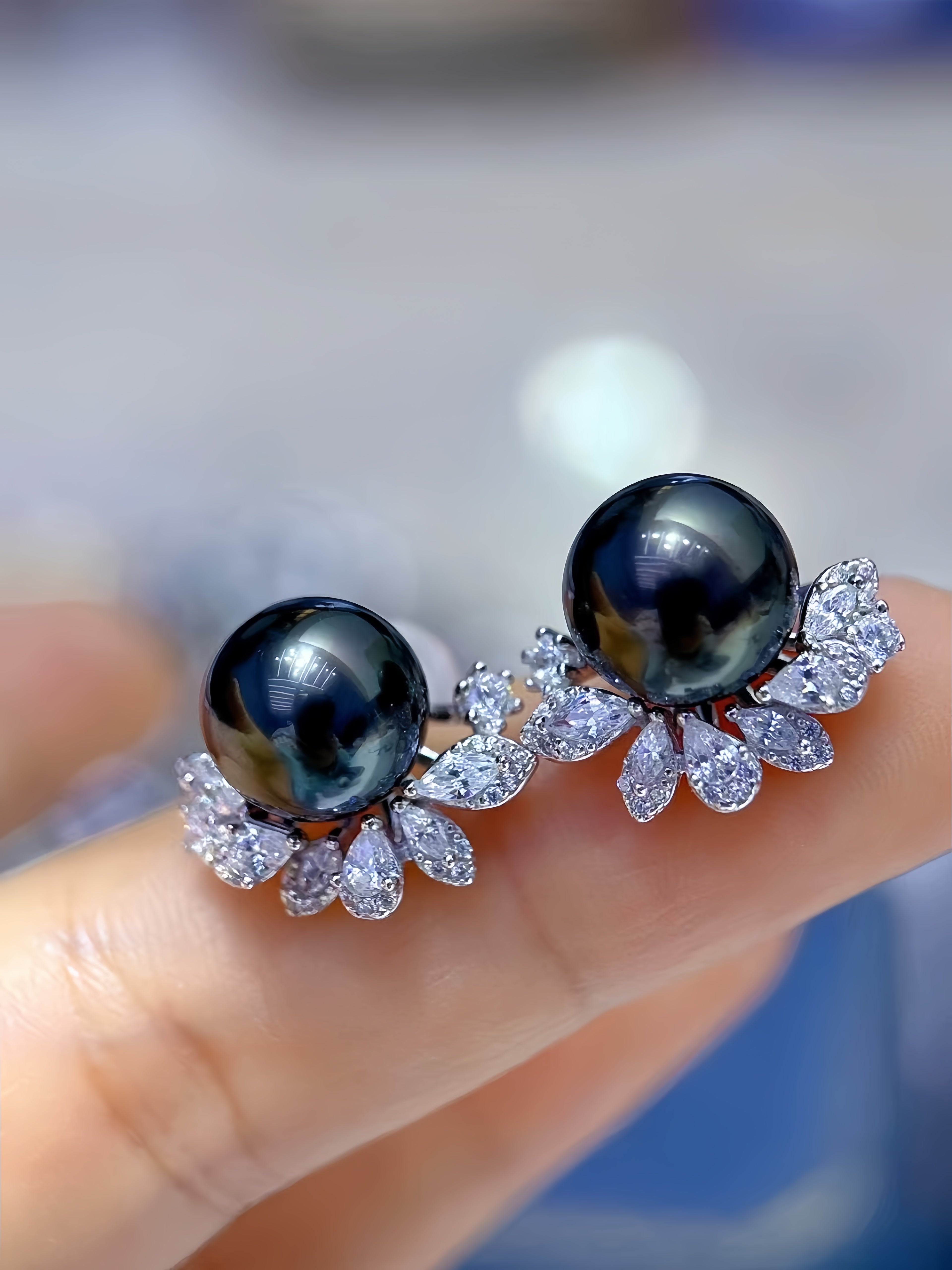 A pair of pearl stud earrings with silver-toned floral settings, held on a person's finger. The round black pearls are flanked by clusters of clear teardrop and round stones.