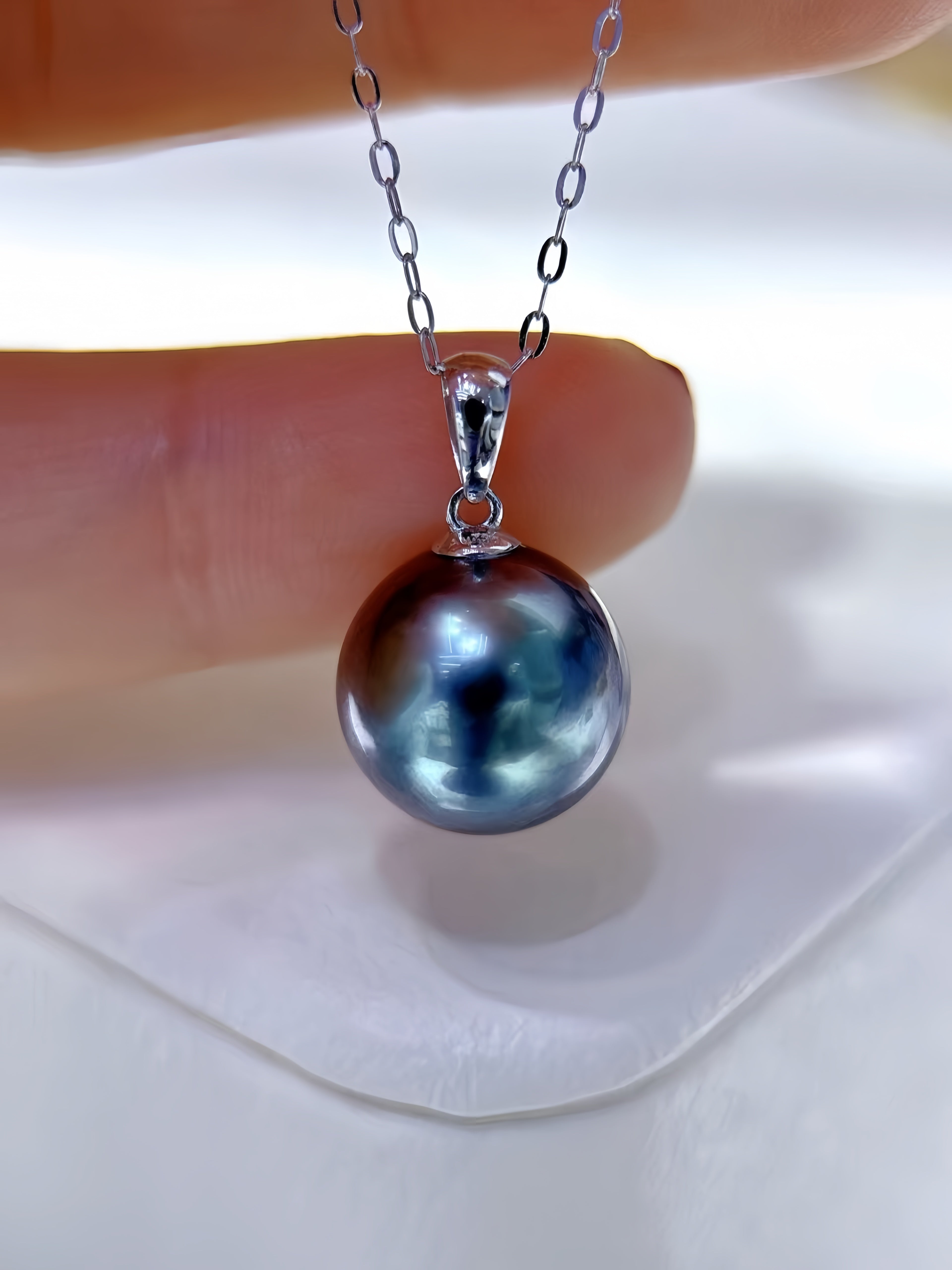A close-up of a silver pendant necklace featuring a large, round, iridescent dark blue-gray pearl, held between fingers against a white background.