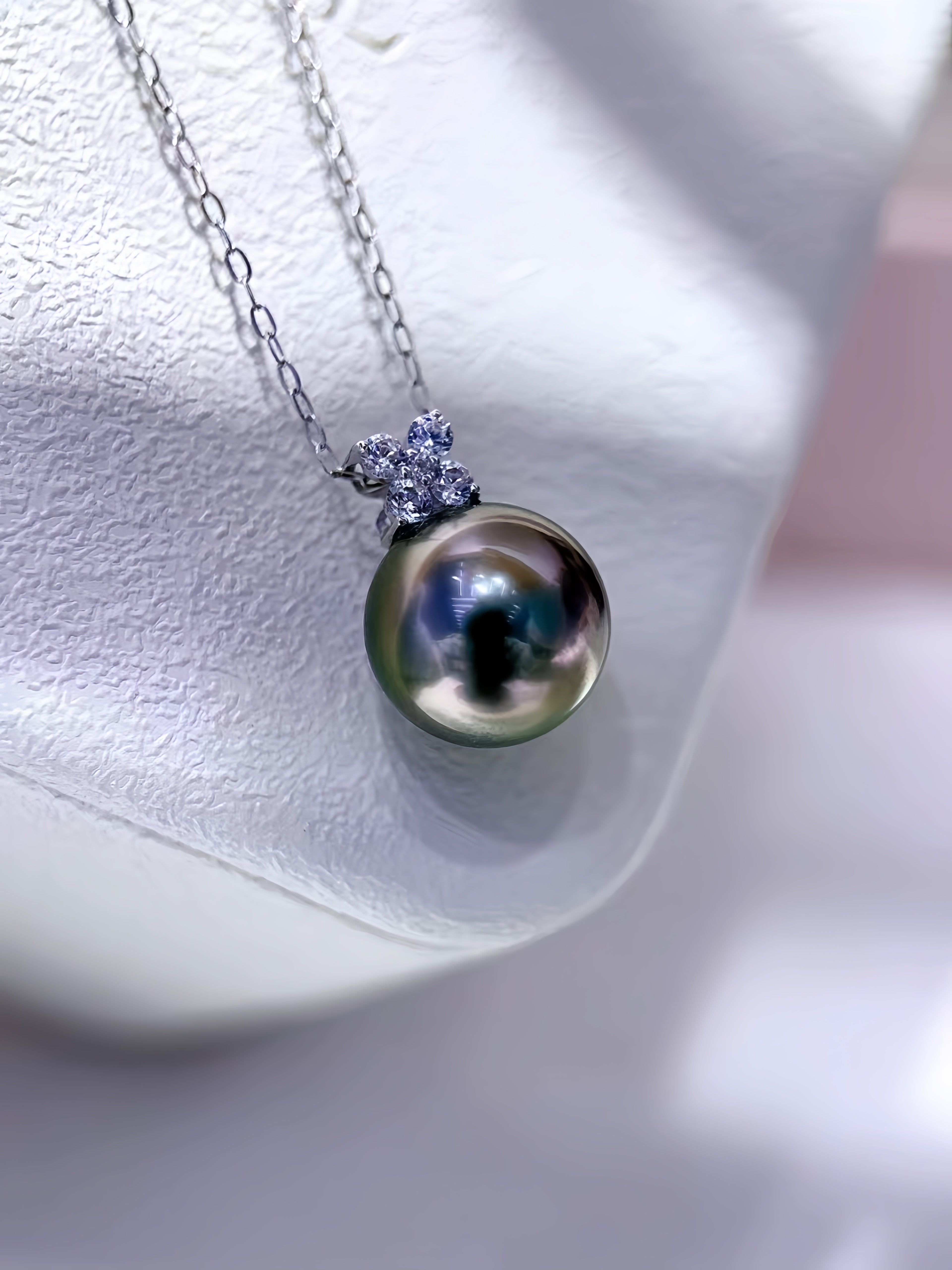 A close-up of a silver chain necklace with a round iridescent pearl pendant and a small cluster of clear stones at the top, displayed on a white jewelry stand.