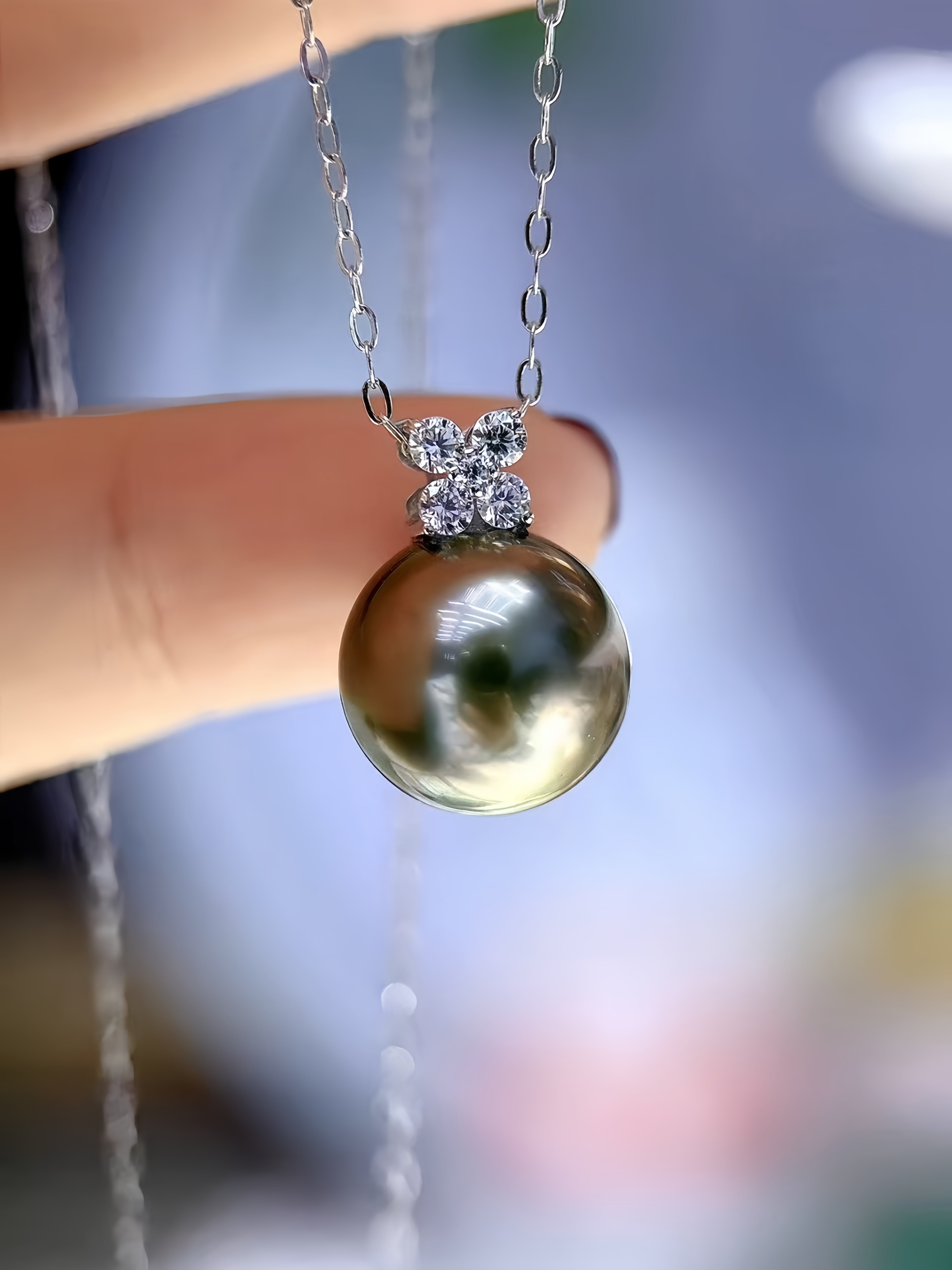 A close-up of a silver chain necklace with a large, glossy dark gold freshwater pearl pendant, held by a person's fingers against a soft, blurred blue background.