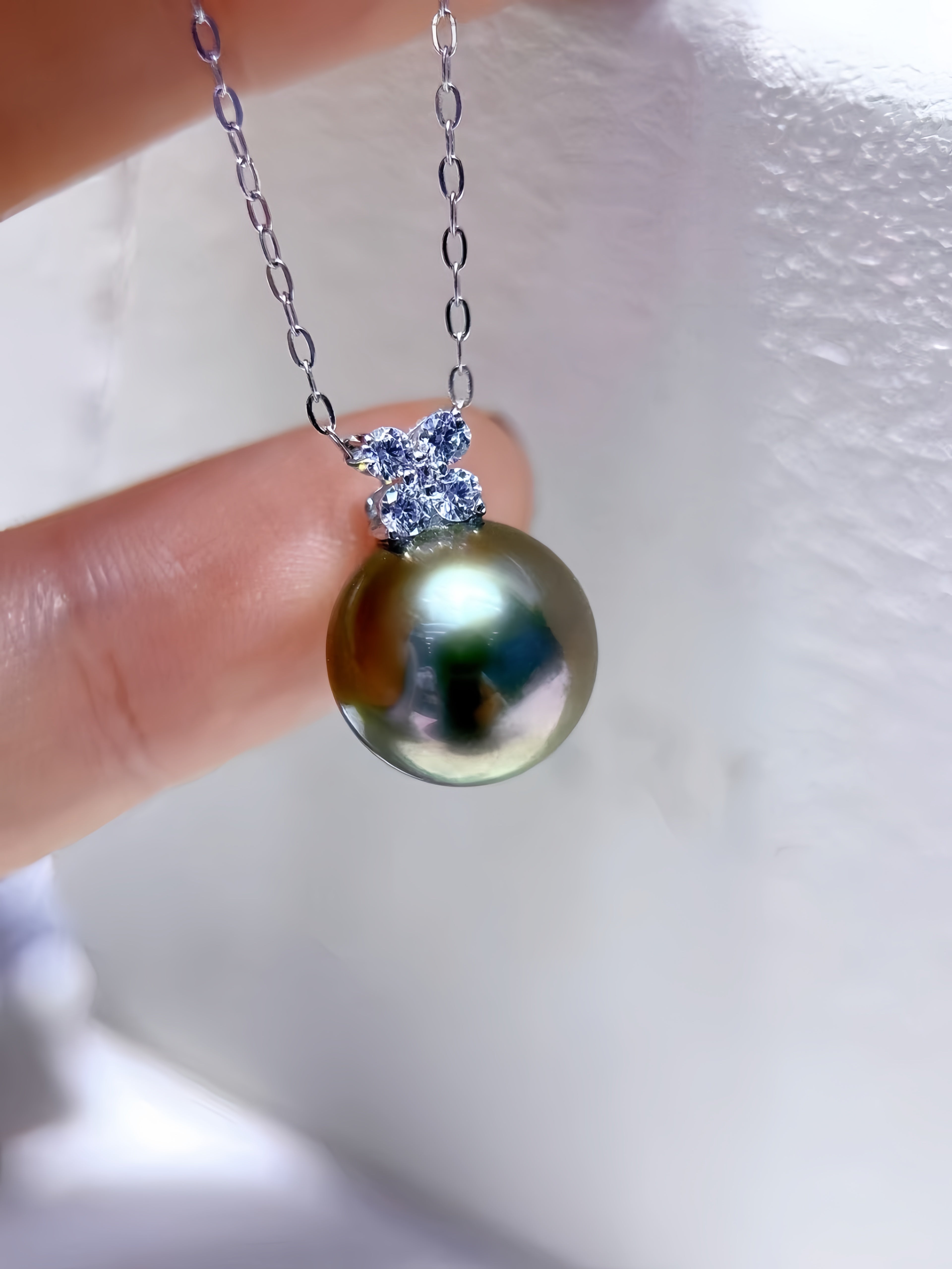 A close-up of a hand holding a silver chain necklace with a large, iridescent green-black pearl pendant and a small cluster of clear stones at the top, set against a soft white textured background.