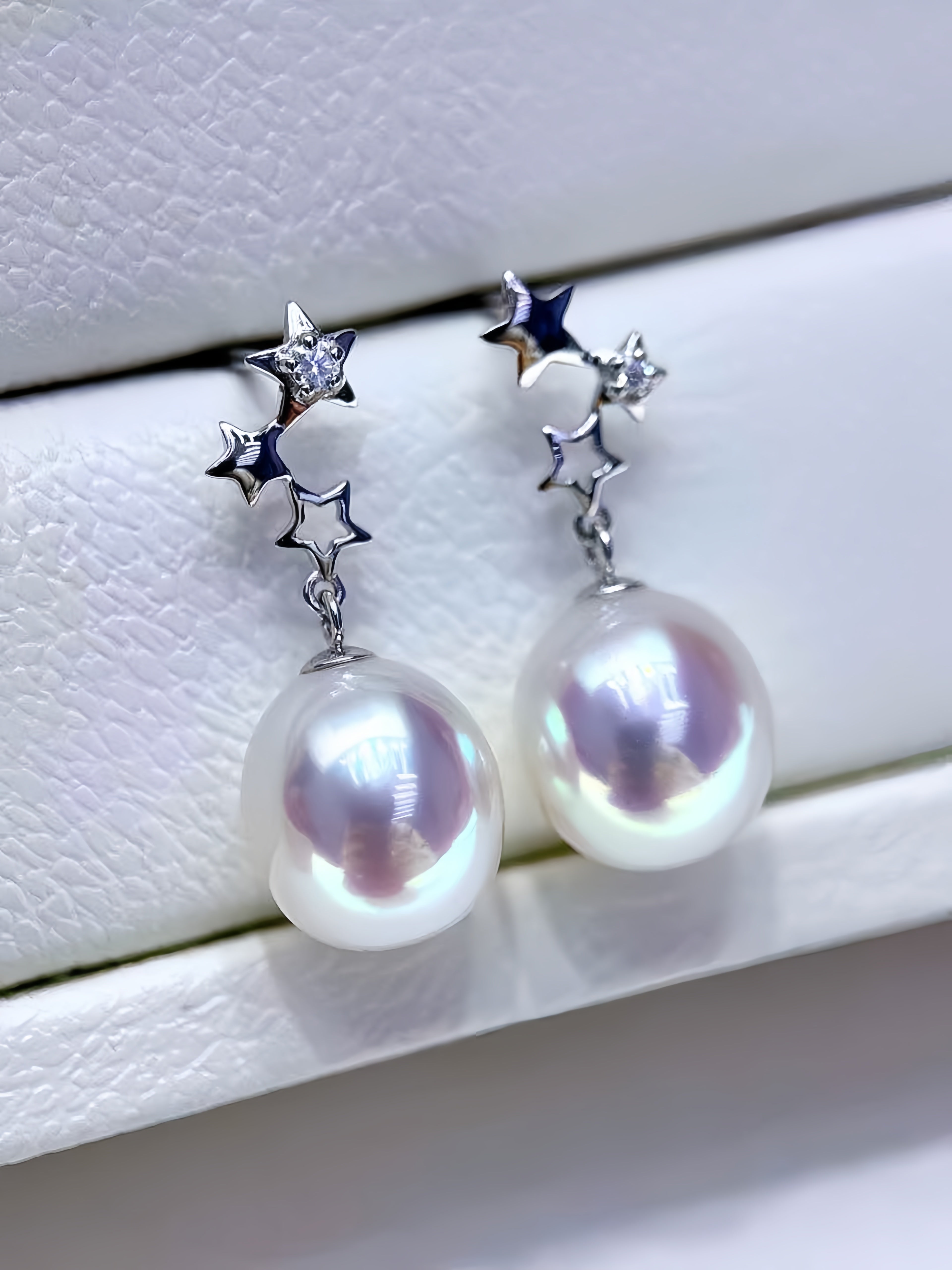 A pair of silver star cluster dangle earrings with large iridescent white freshwater pearls, displayed on a textured white jewelry box.
