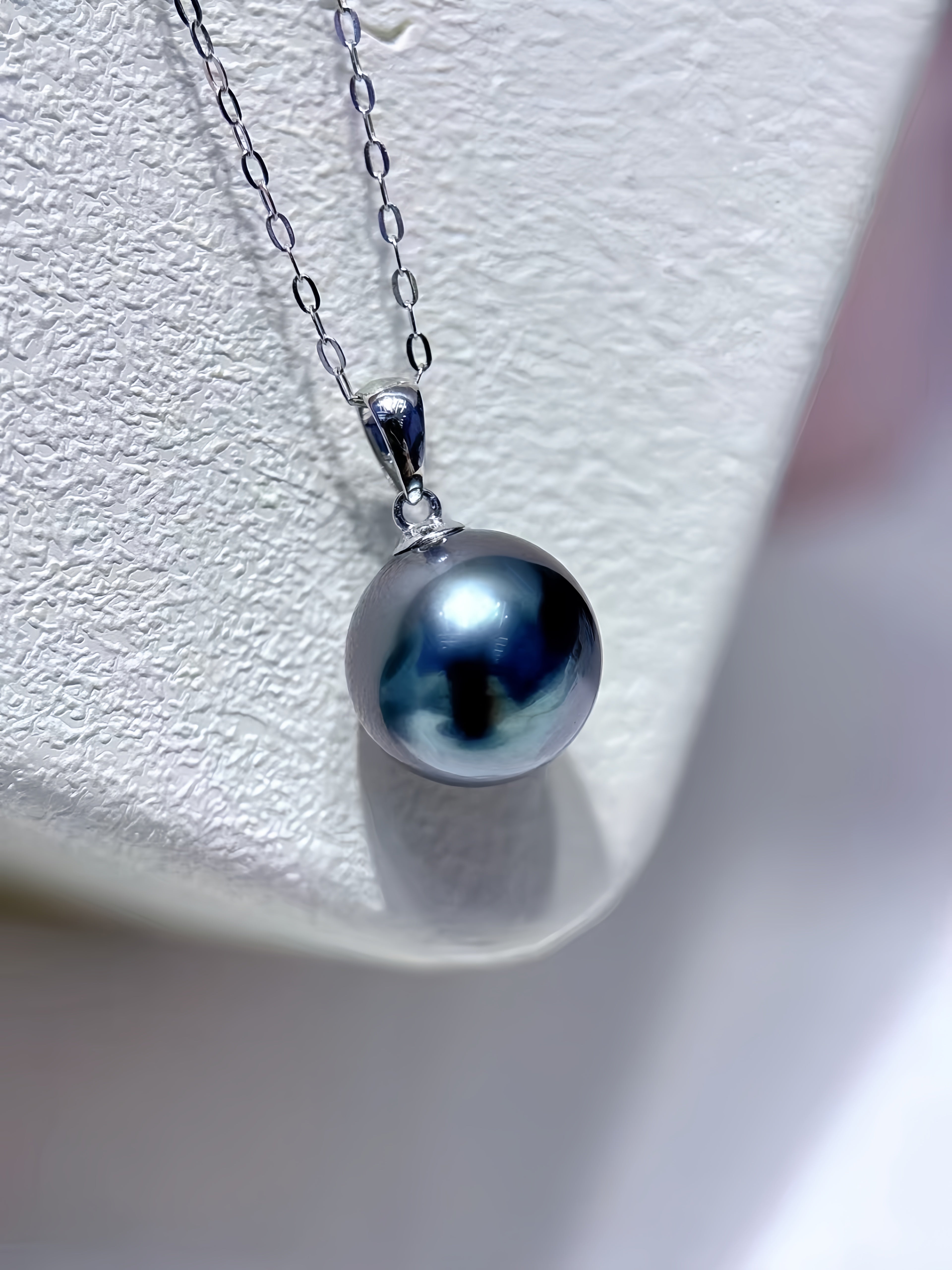 A silver pendant necklace featuring a single round, dark blue-gray pearl on a delicate silver chain, displayed on a textured white jewelry stand.