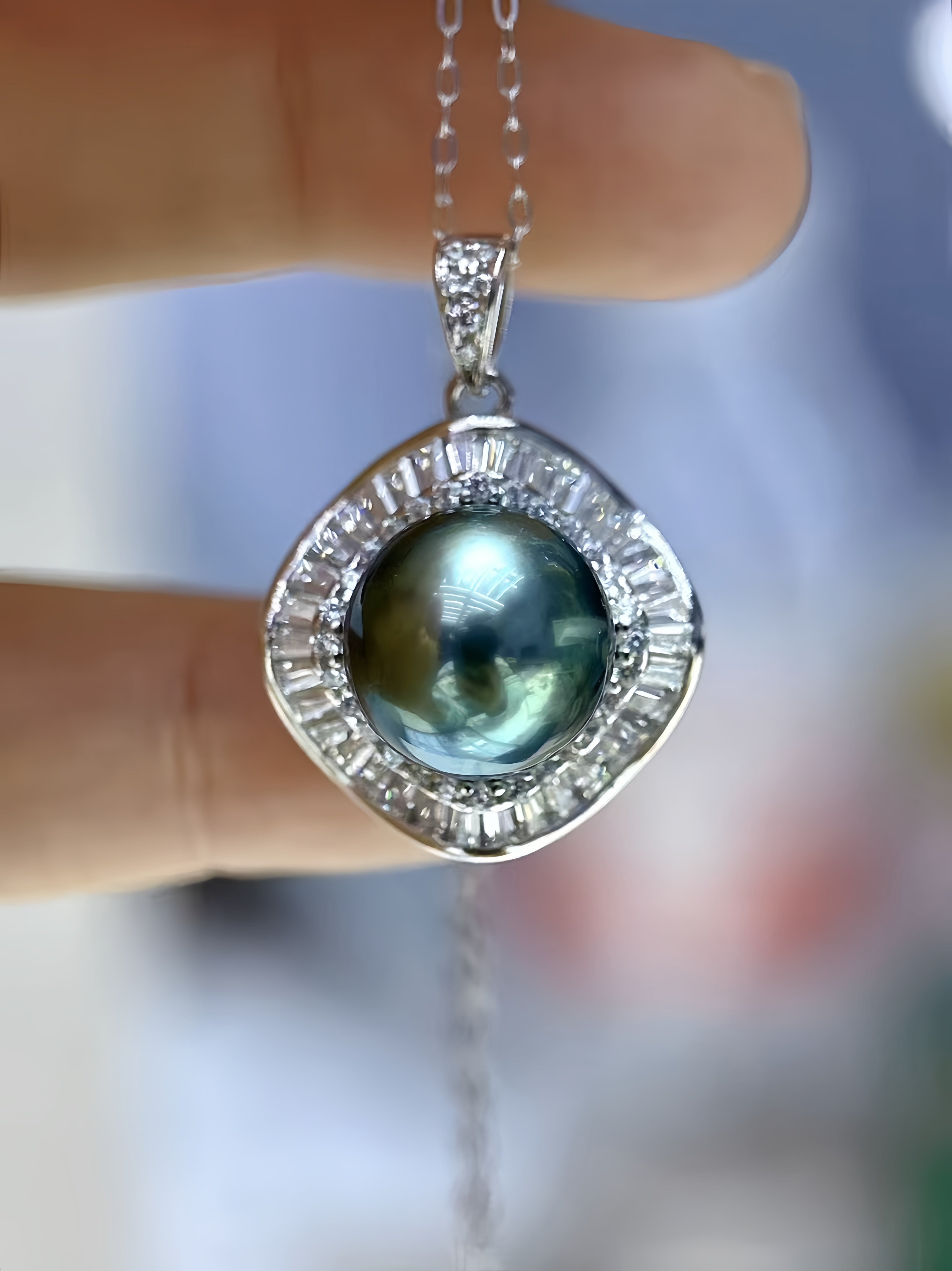 A close-up of a silver pendant necklace held between fingers. The pendant features a central iridescent green-blue pearl surrounded by a halo of small clear stones, with a larger bezel-set pearl at the top of the pendant.