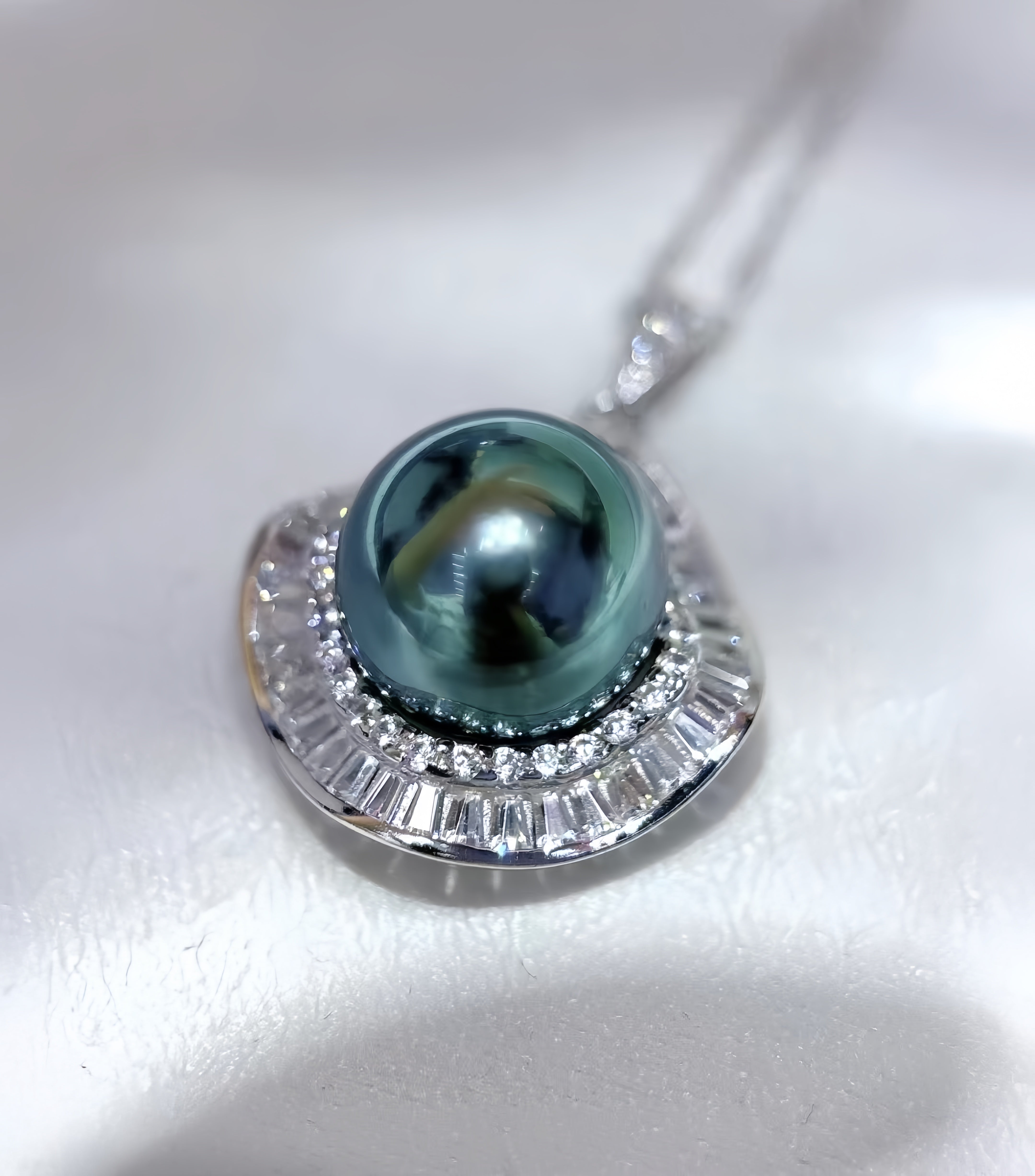 A close-up of a pearl pendant necklace featuring a dark green teardrop-shaped pearl set in a silver-toned halo with clear baguette and round stones, resting on a white textured surface.