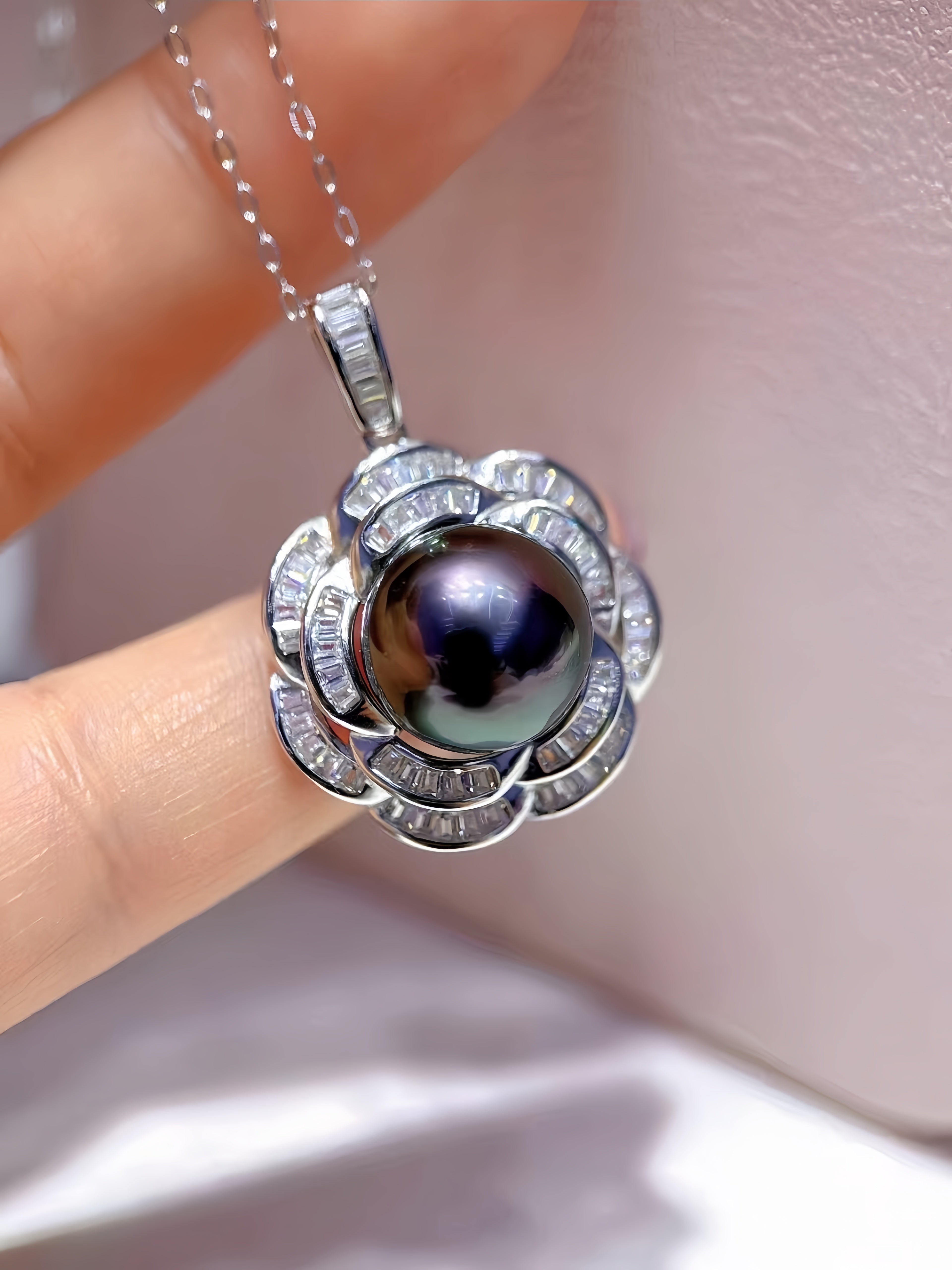 A close-up of a silver pendant necklace with a dark, iridescent pearl surrounded by small clear stones, held between fingers against a soft pink background.