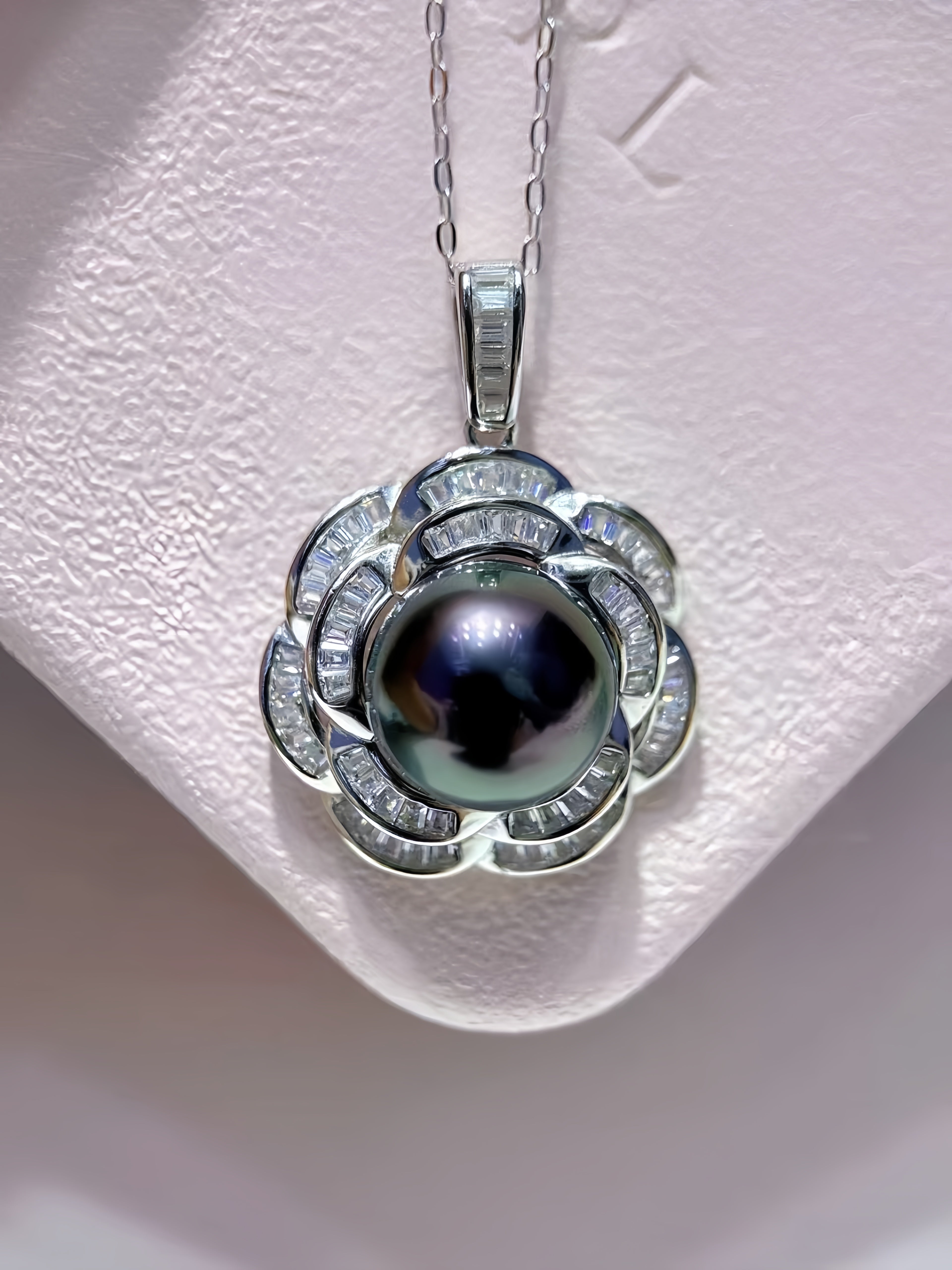 A silver pendant necklace featuring a central dark iridescent pearl surrounded by silver petal-shaped settings set with small clear stones, displayed on a textured white jewelry stand.