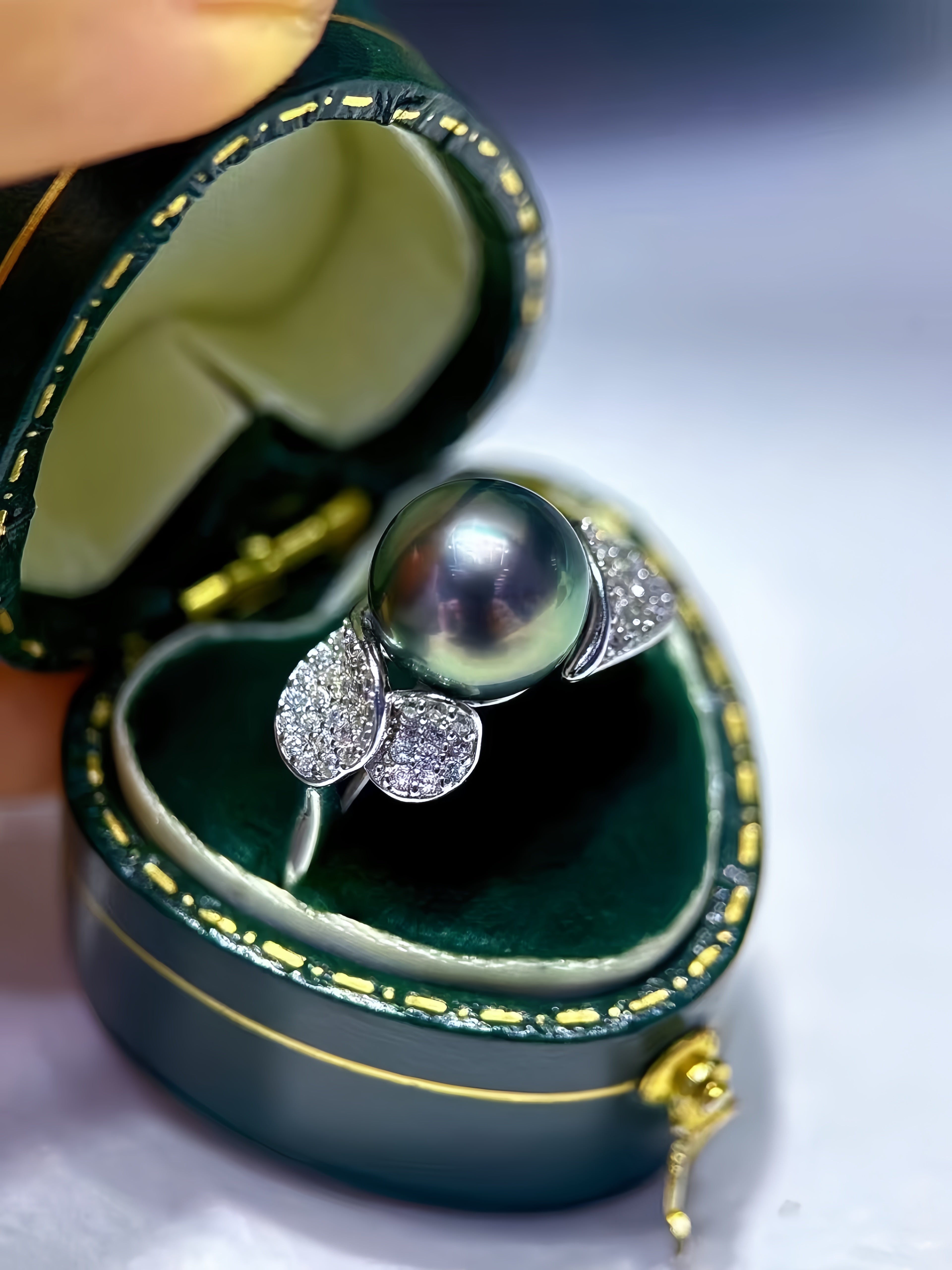 A close-up of a ring displayed inside an open dark green jewelry box. The ring features a large round dark pearl on a silver-toned setting with small pavé-set clear stones.