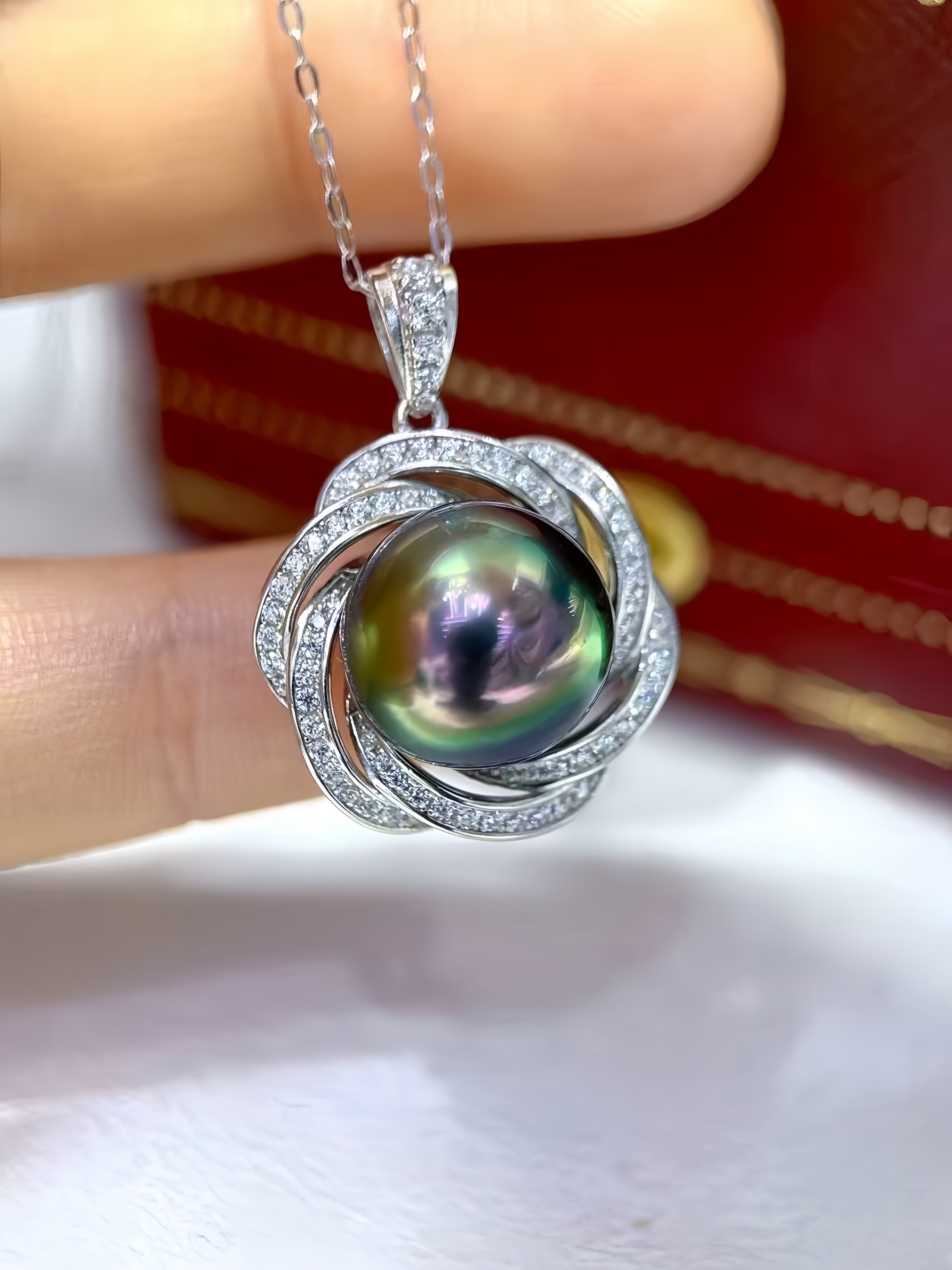 A close-up of a silver pendant necklace with a round iridescent pearl surrounded by silver filigree and small clear stones, held between fingers against a red and white background.