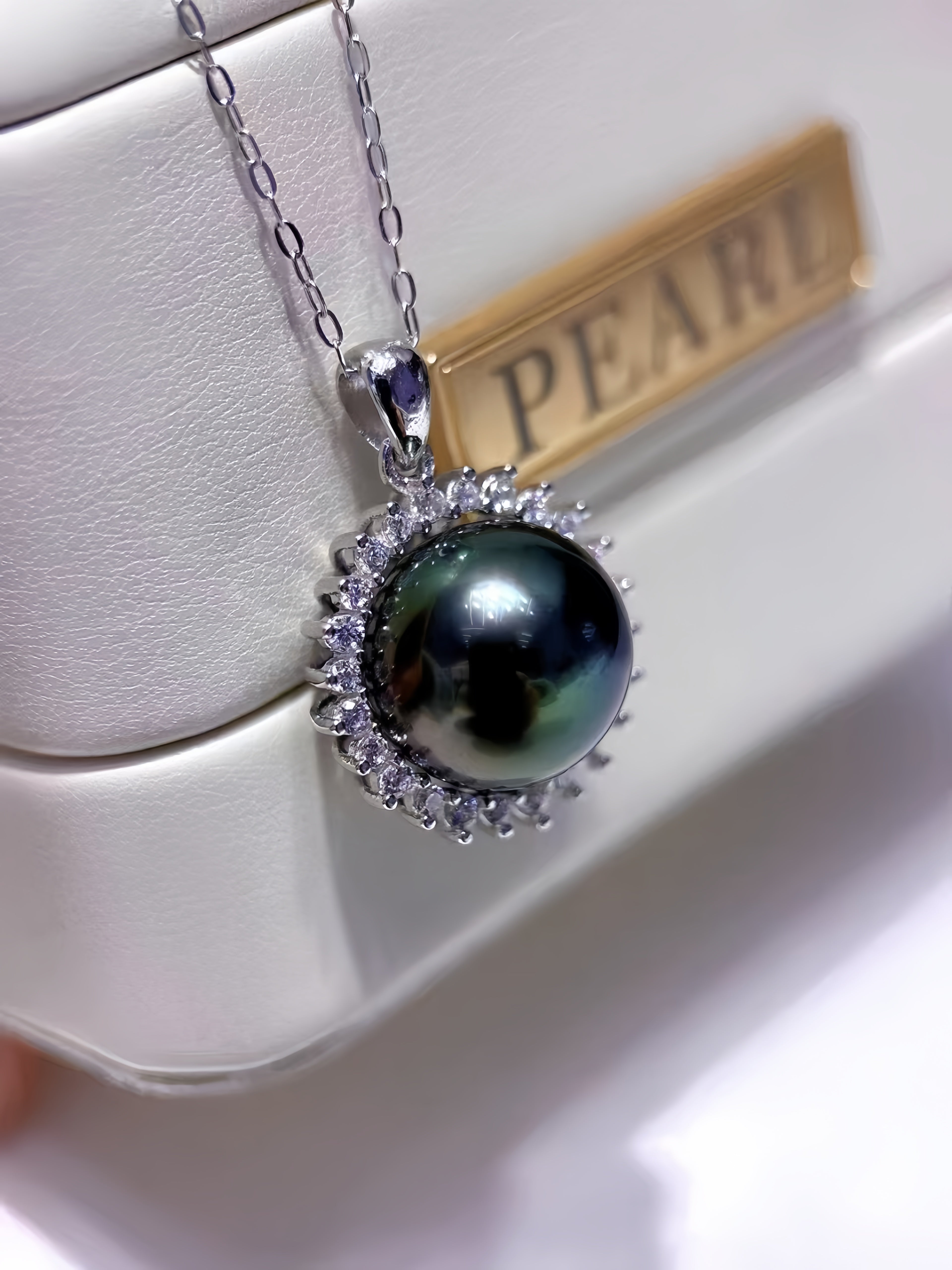 A silver pendant necklace featuring a large, dark green-black pearl surrounded by a halo of small clear stones, resting on a white jewelry box with a gold 'PEARL' label.