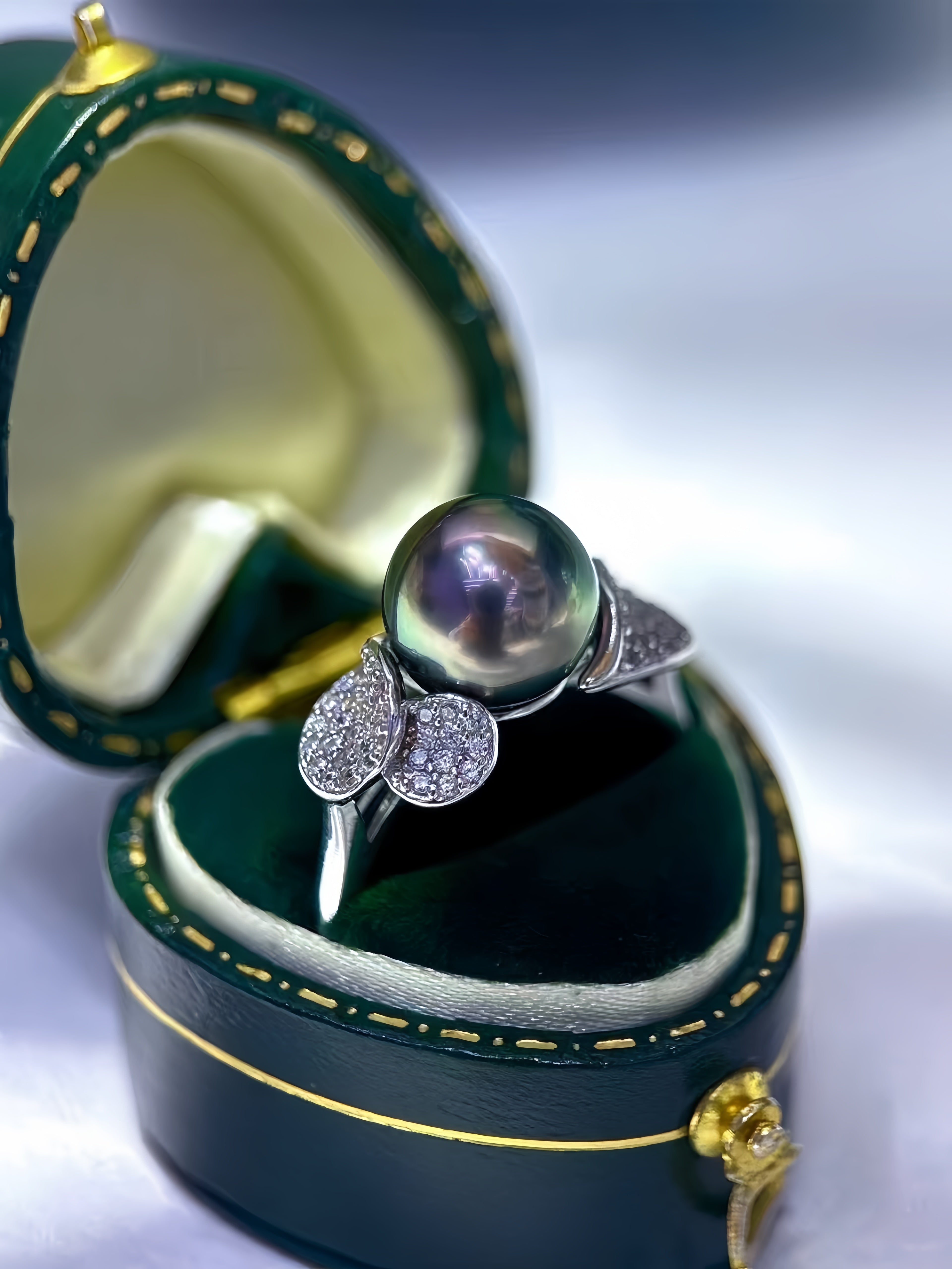 A close-up of a pearl ring displayed in an open dark green jewelry box. The ring features a large round iridescent pearl set in a silver-toned band with two diamond-accented butterfly motifs on either side.