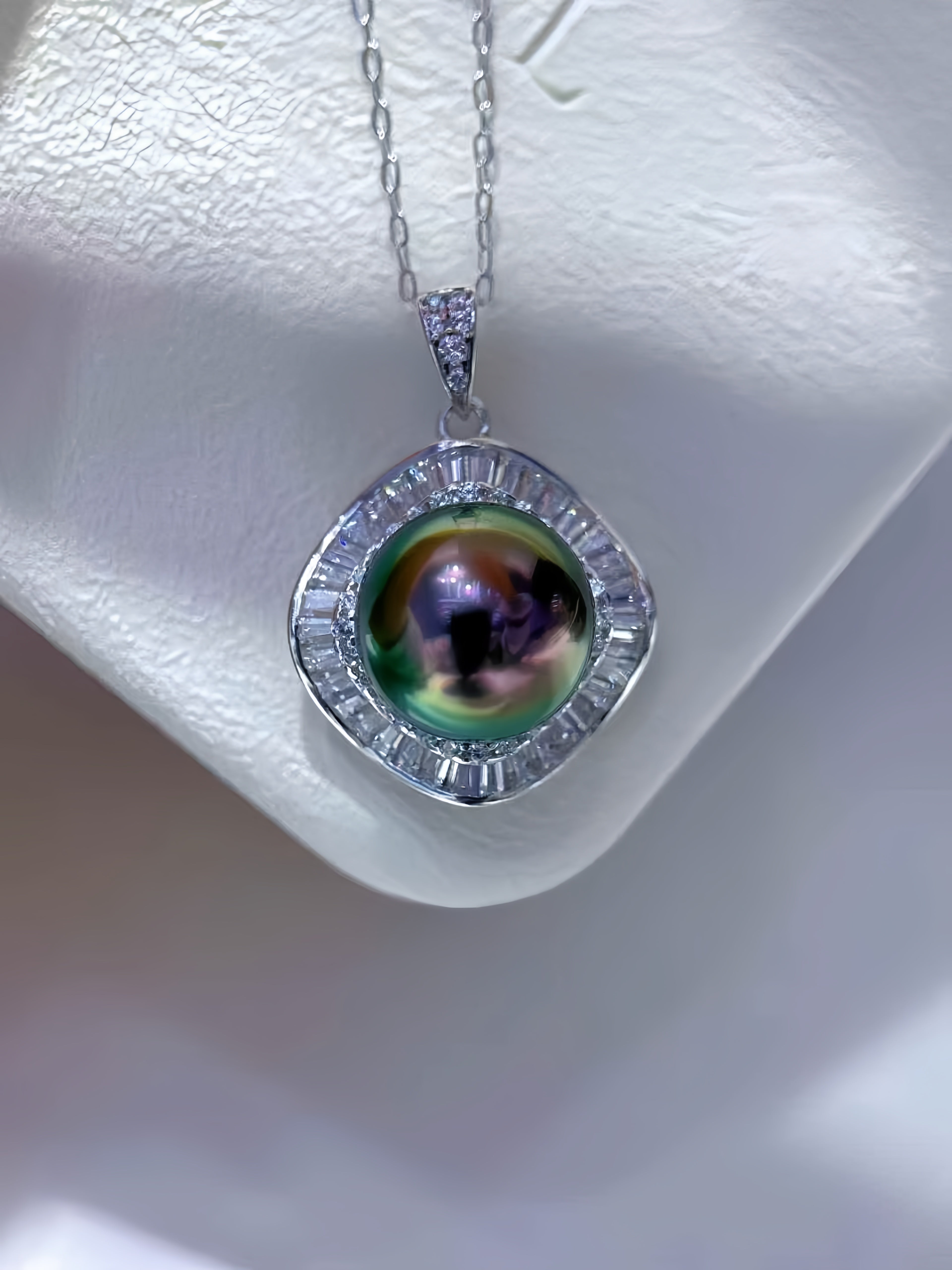 A silver pendant necklace featuring a large, iridescent green pearl surrounded by a halo of small clear stones, displayed on a white jewelry bust.