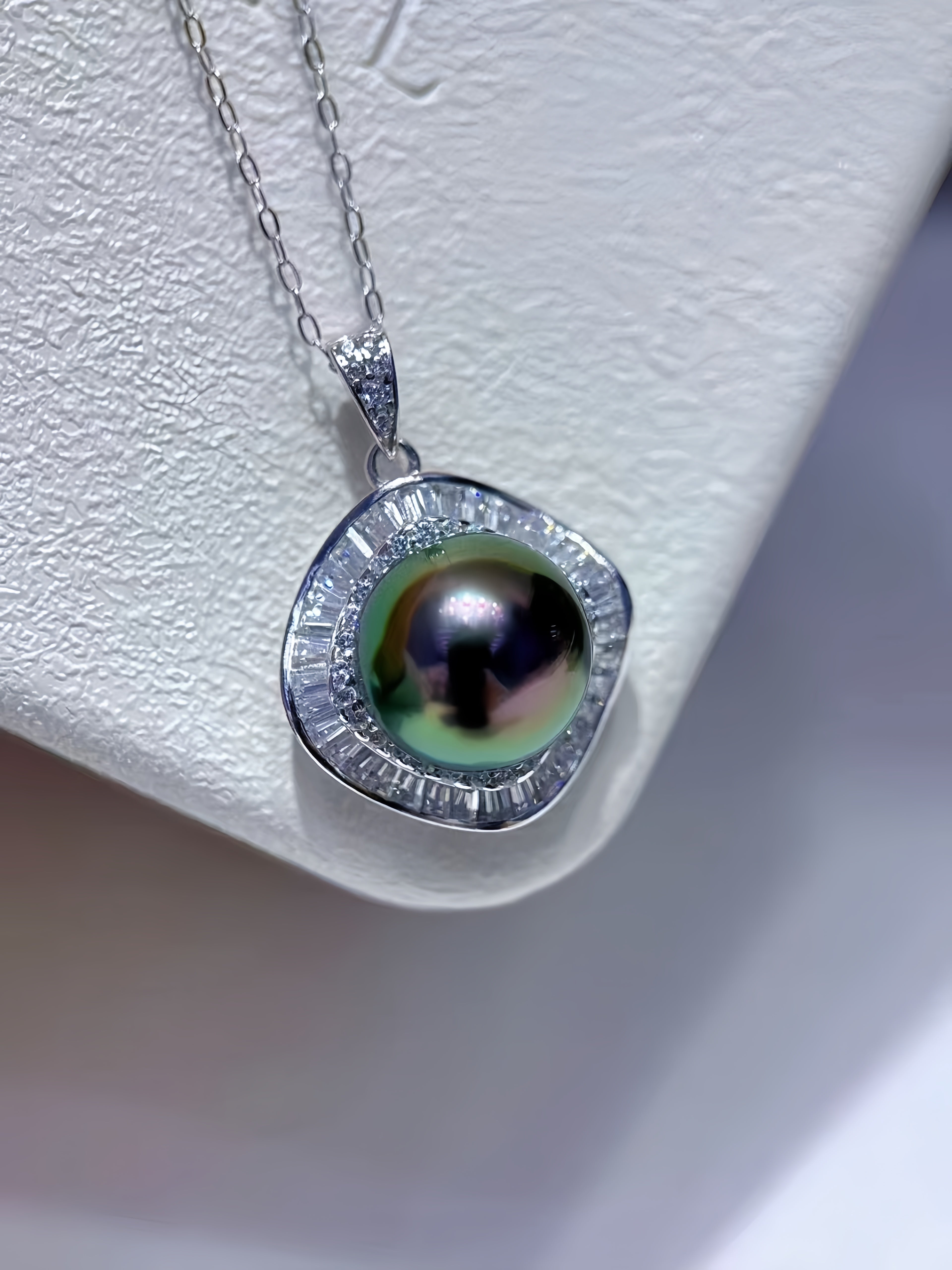 A silver pendant necklace featuring a round iridescent green pearl surrounded by a halo of small clear stones, displayed on a textured white jewelry stand.