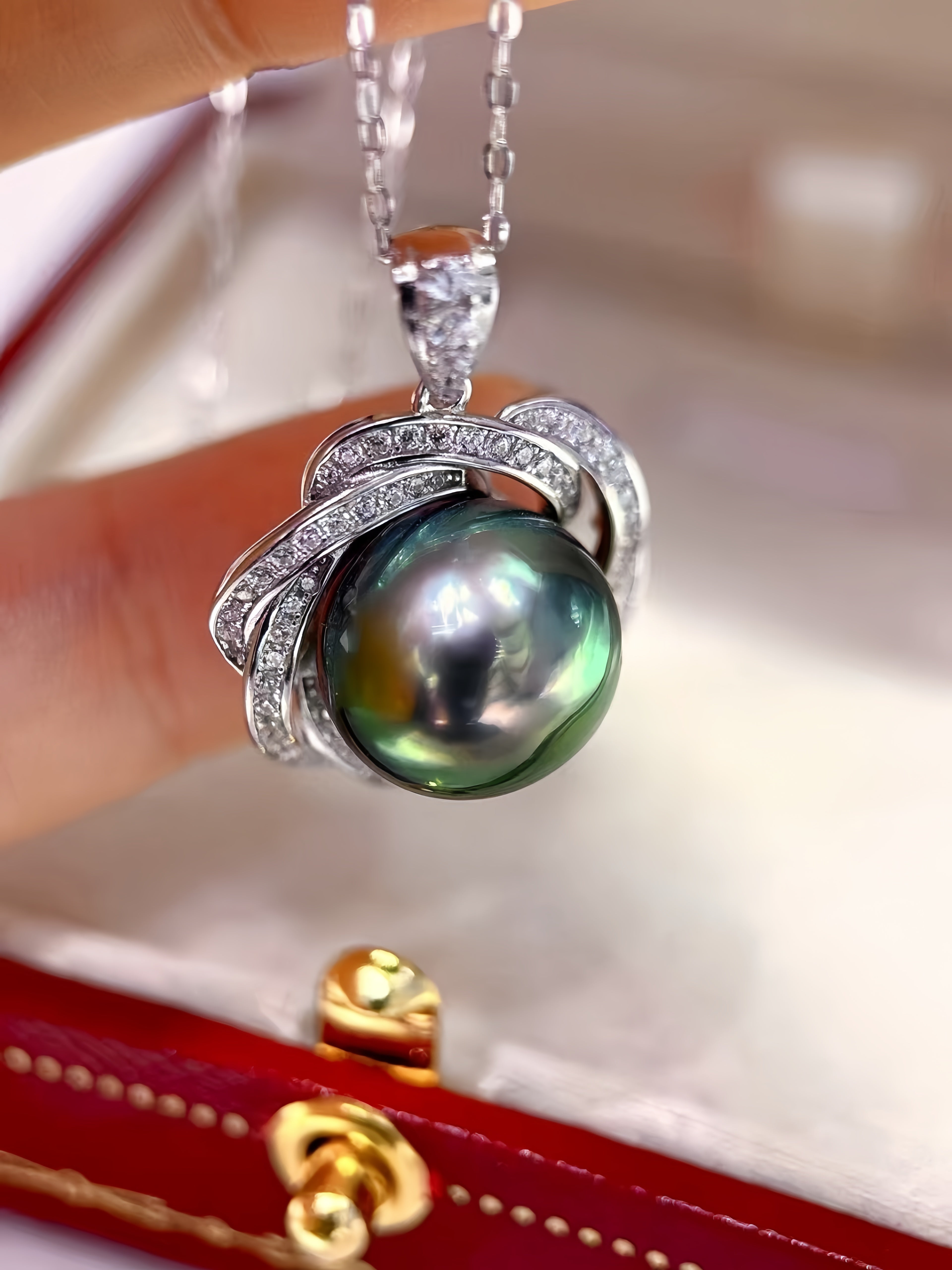 A close-up of a silver pendant necklace featuring a large, round, iridescent green pearl surrounded by silver curved bands set with small clear stones, held by fingers above an open red jewelry box.