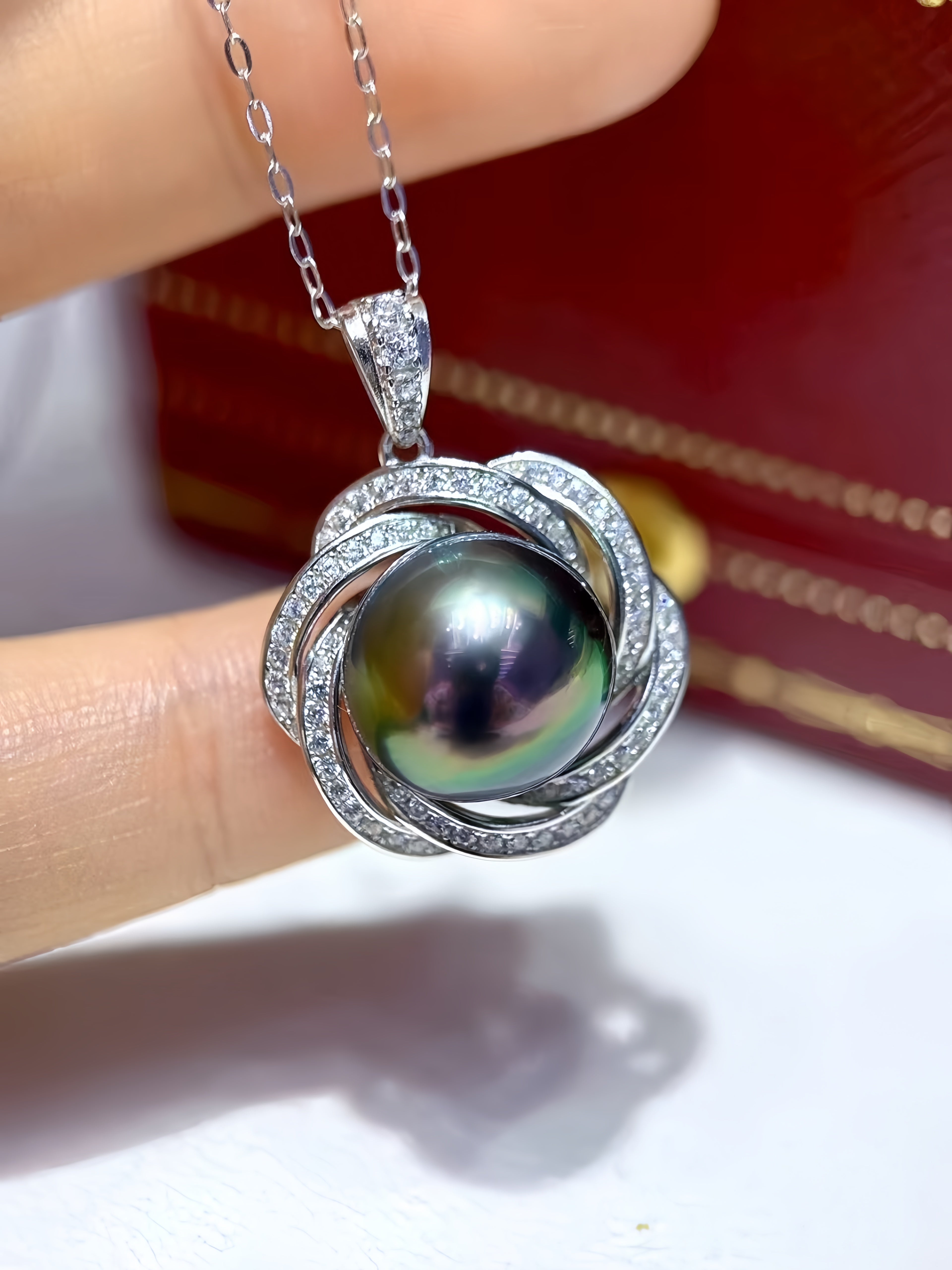 A close-up of a silver pendant necklace with a large iridescent freshwater pearl and small clear stones, held between fingers with a red jewelry box blurred in the background.