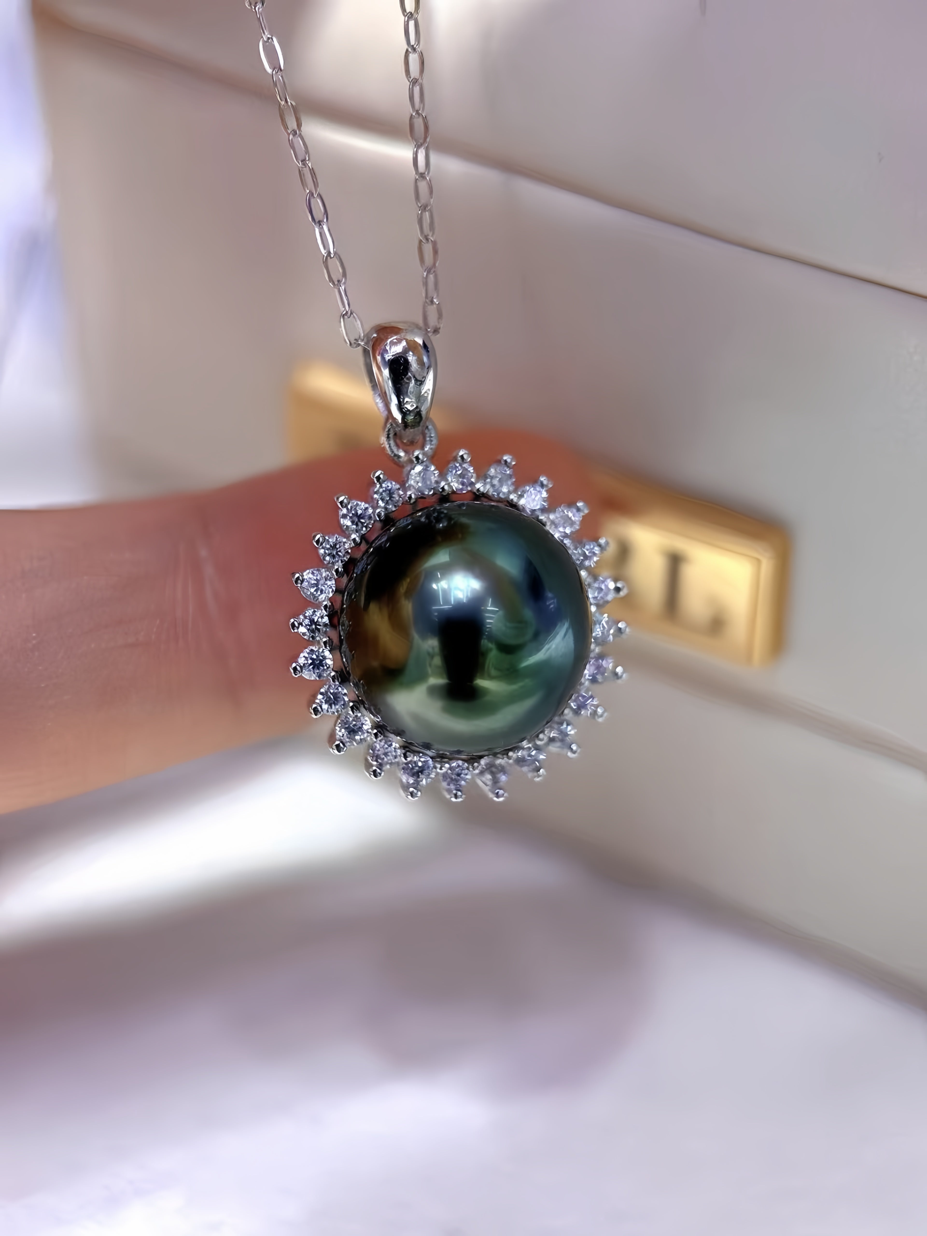 A silver pendant necklace featuring a large, round, dark green-black pearl surrounded by a halo of small clear stones, held by a hand against a light-colored jewelry box background.