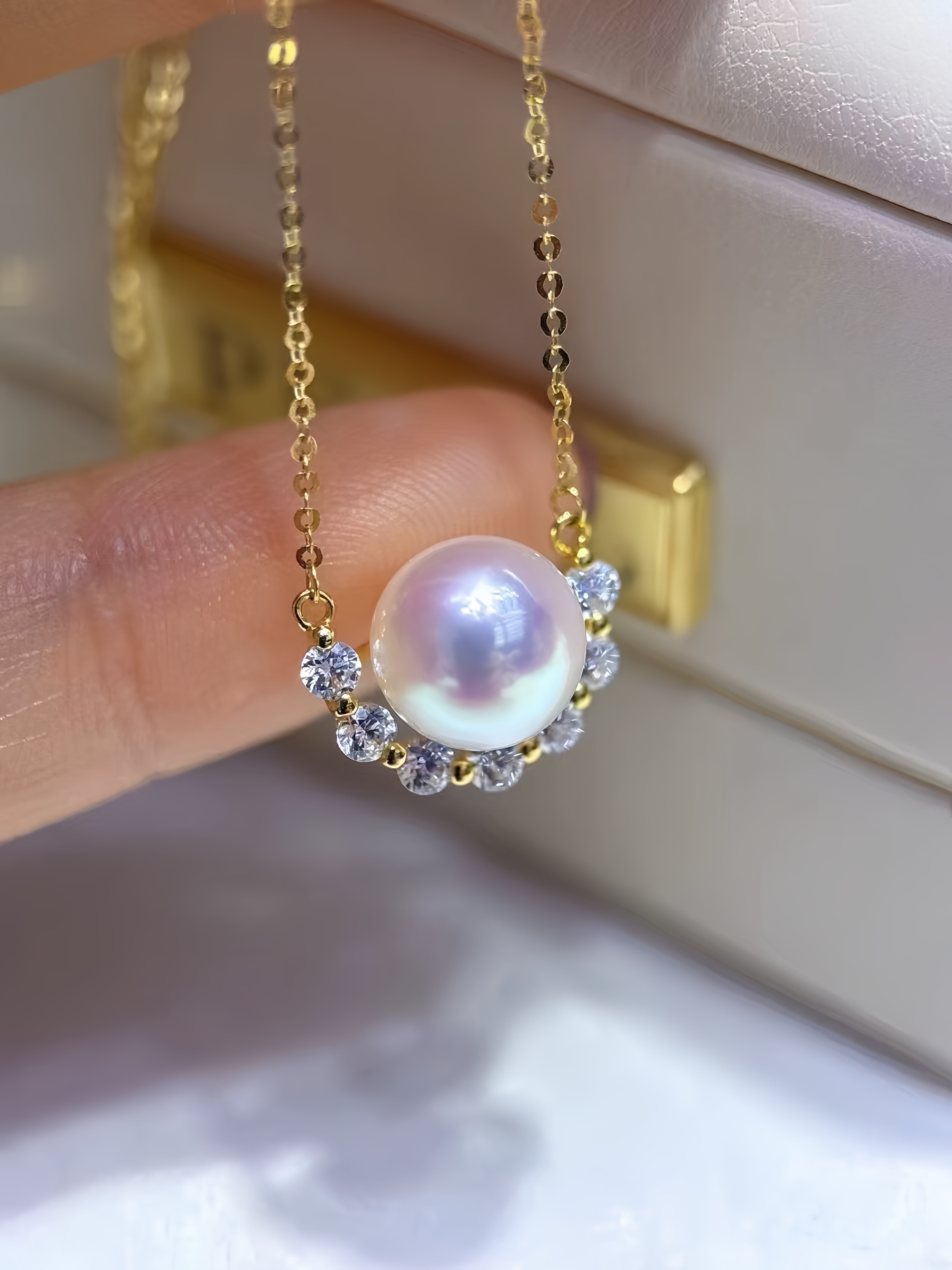 A gold chain necklace with a large lustrous pearl pendant surrounded by small clear stones, held by a finger in front of a light jewelry box.