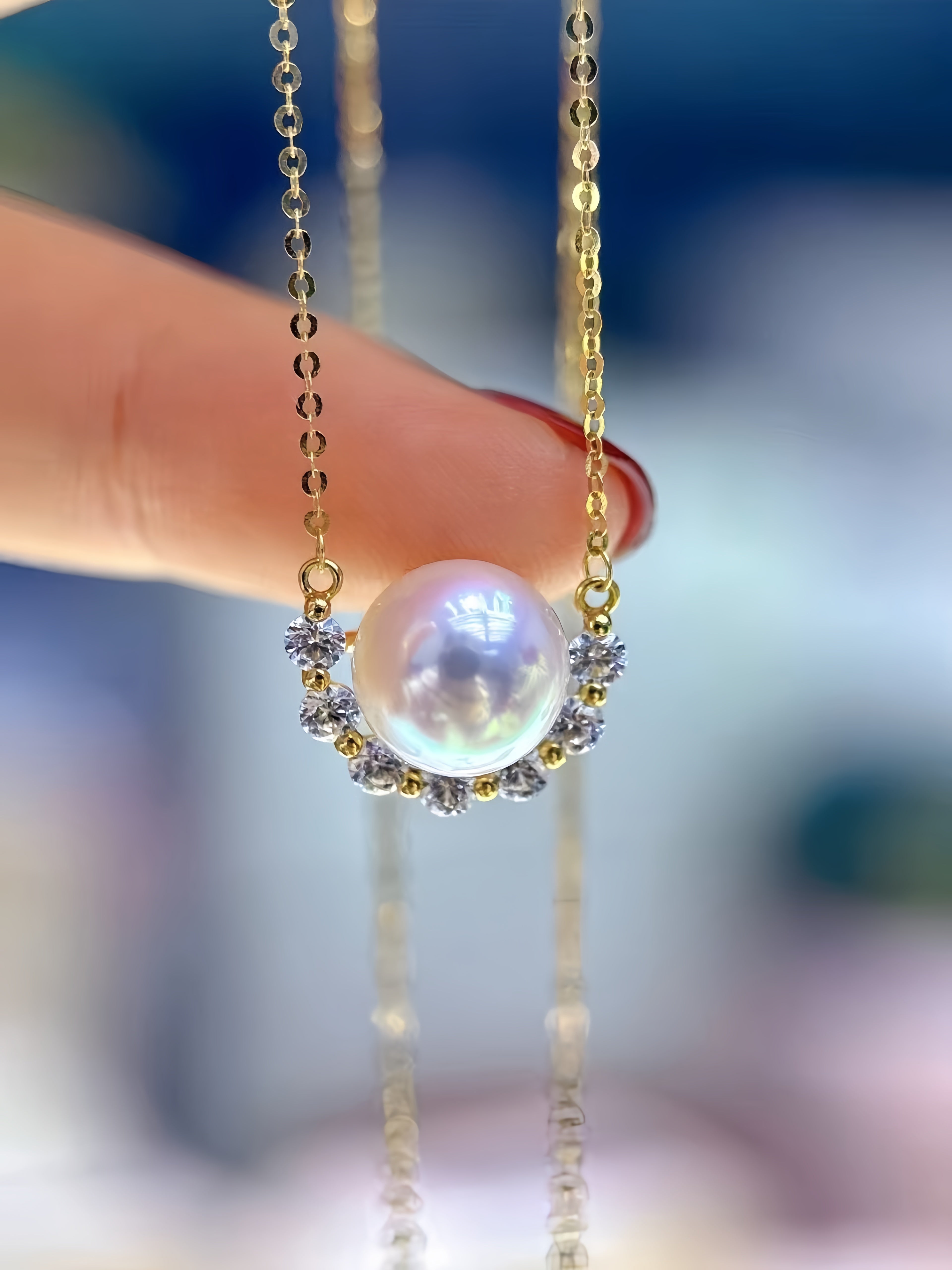 A gold necklace with a large iridescent white pearl pendant, accented by small clear stones, held by a person's finger against a blurred blue background.