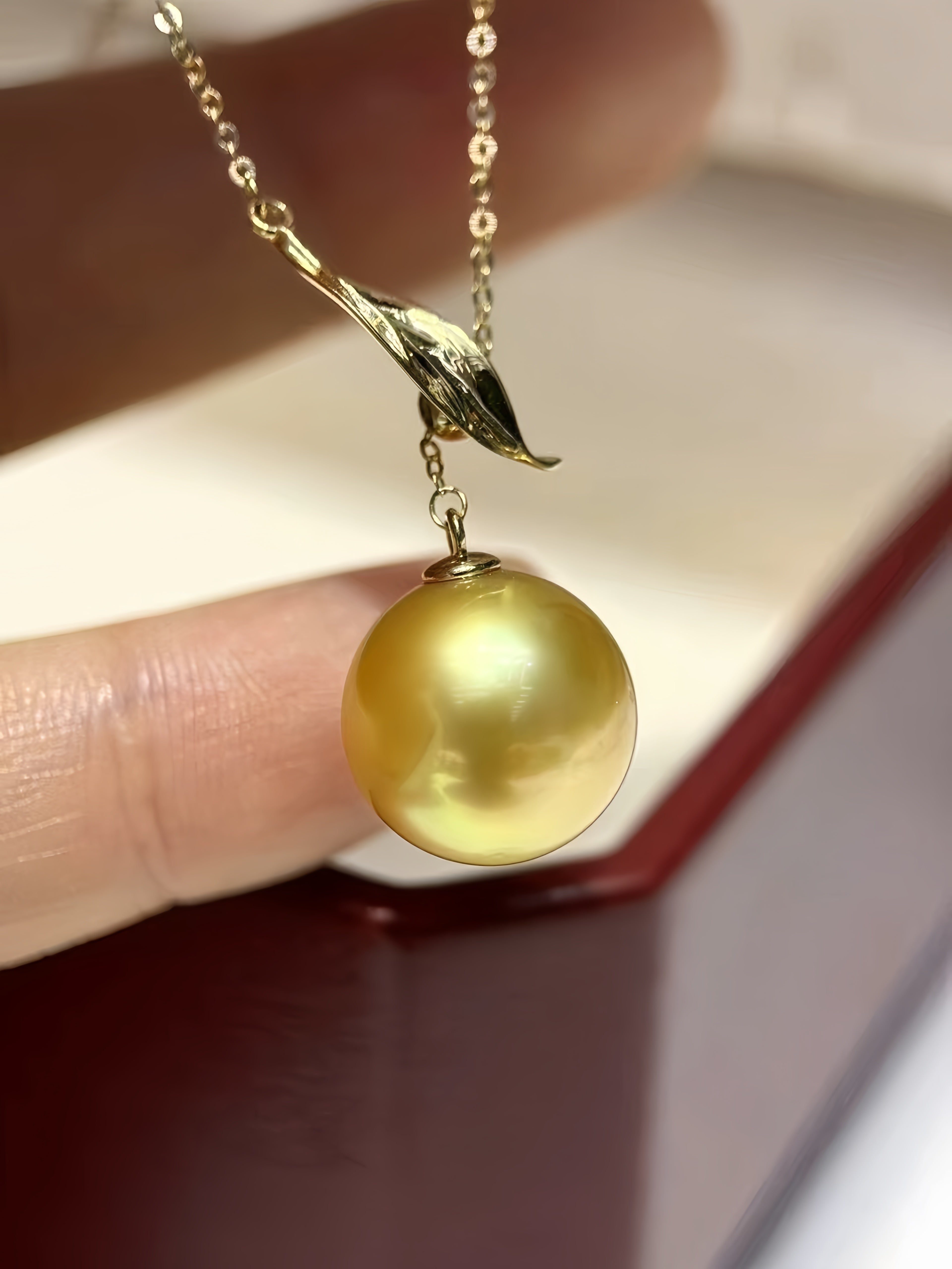 A close-up of a gold pendant necklace featuring a large, lustrous golden pearl and a small gold leaf-shaped charm, held by fingers next to a dark red jewelry box.