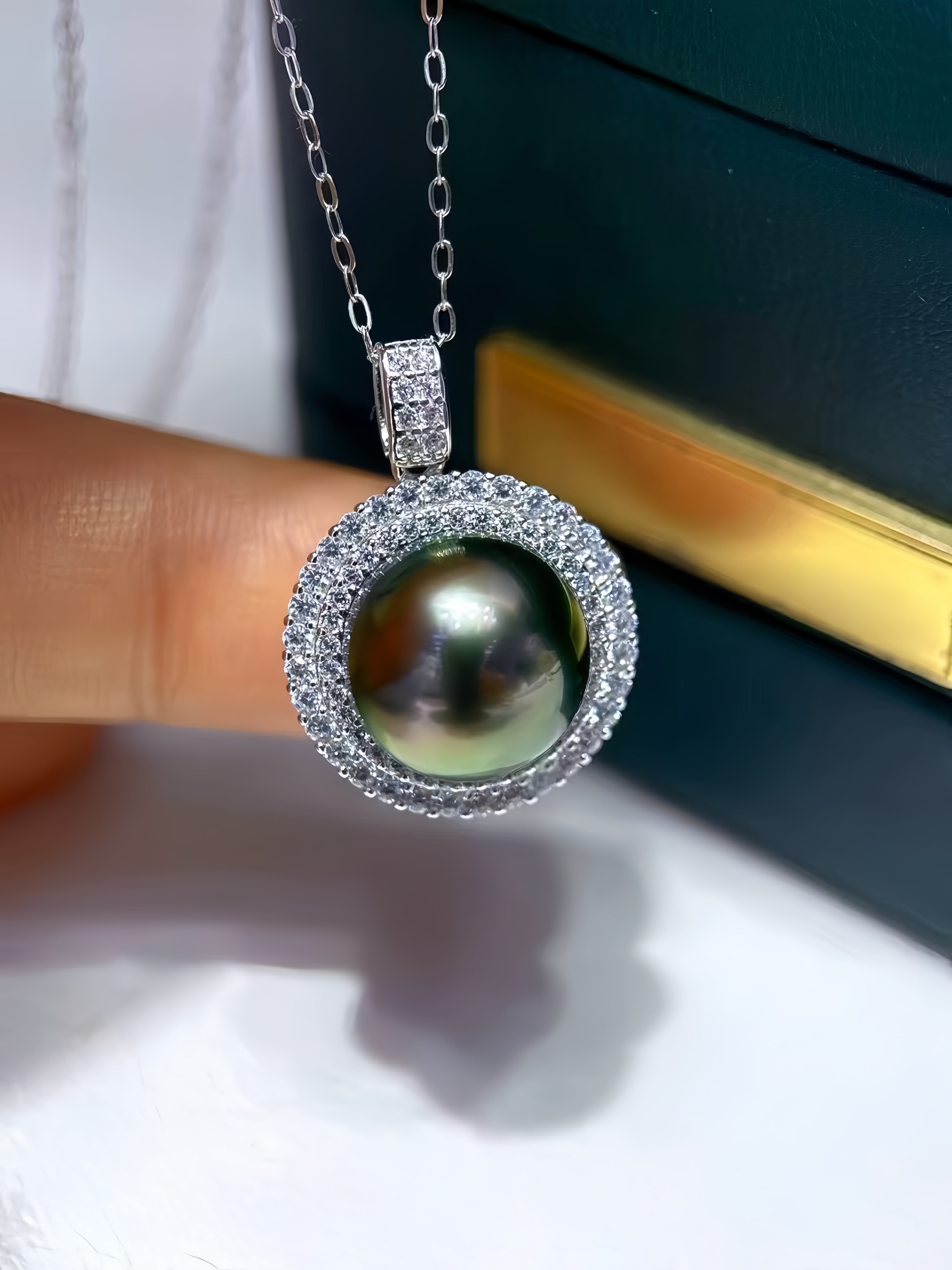 A close-up of a silver pendant necklace featuring a large round green-black pearl surrounded by a halo of small clear stones, held by a finger against a dark green jewelry box.