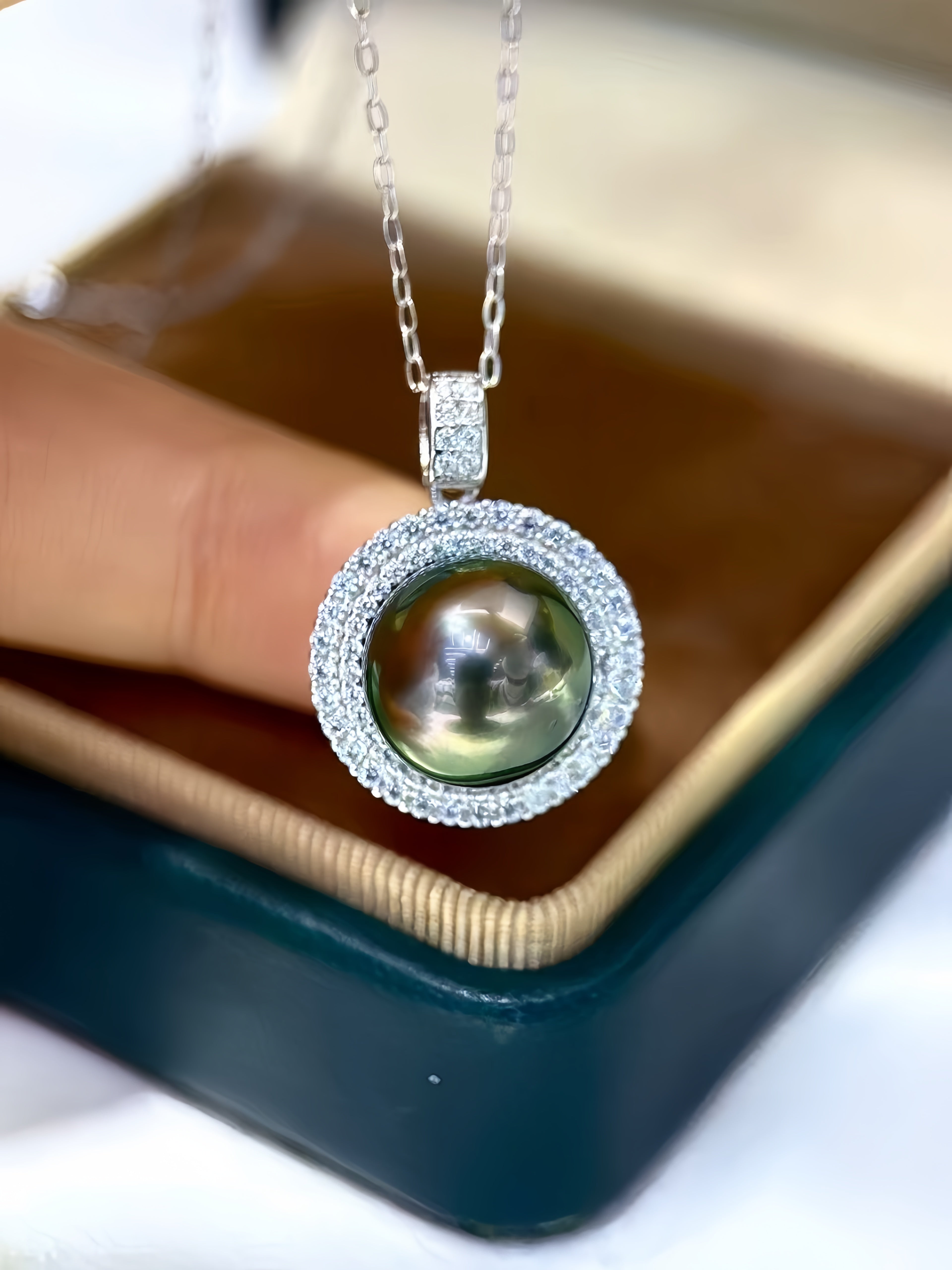 A silver pendant necklace with a large iridescent green pearl surrounded by a halo of small clear stones, held by a hand over an open dark teal jewelry box.