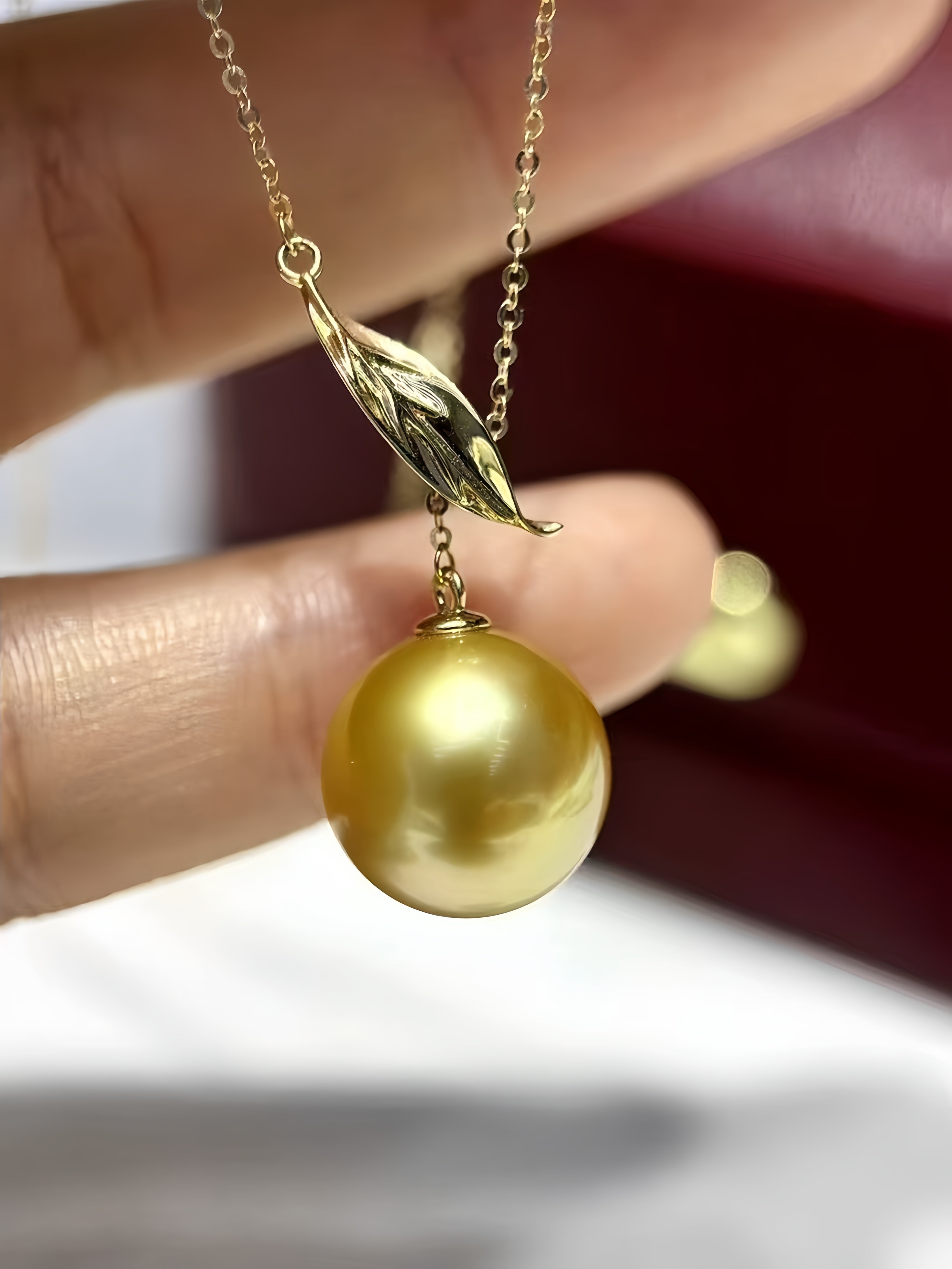 A gold pendant necklace featuring a large, lustrous gold pearl and a leaf-shaped charm, held by a hand against a blurred dark red and white background.