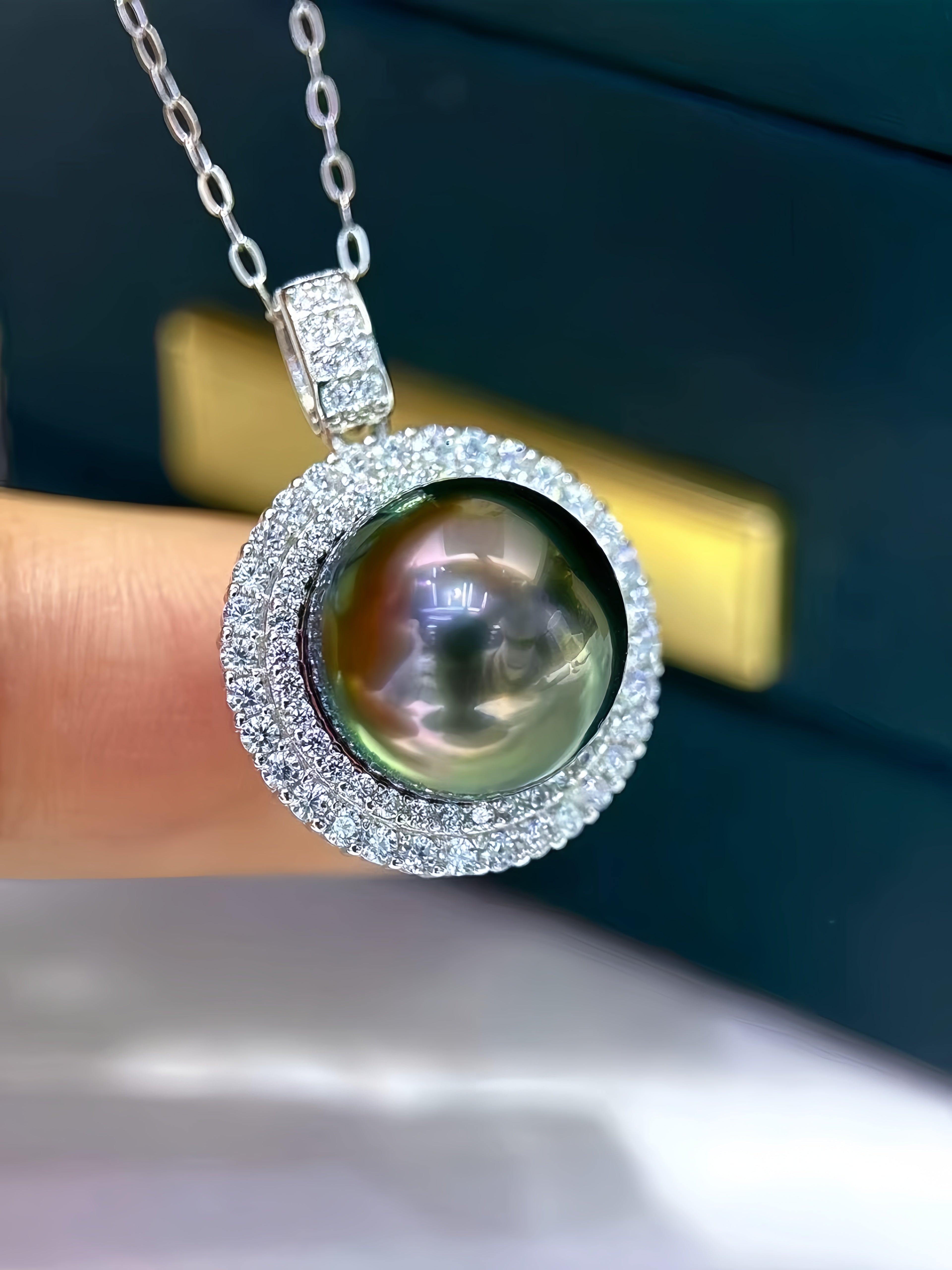 A close-up of a round freshwater pearl pendant necklace with a silver halo and chain, held by a finger against a dark background.