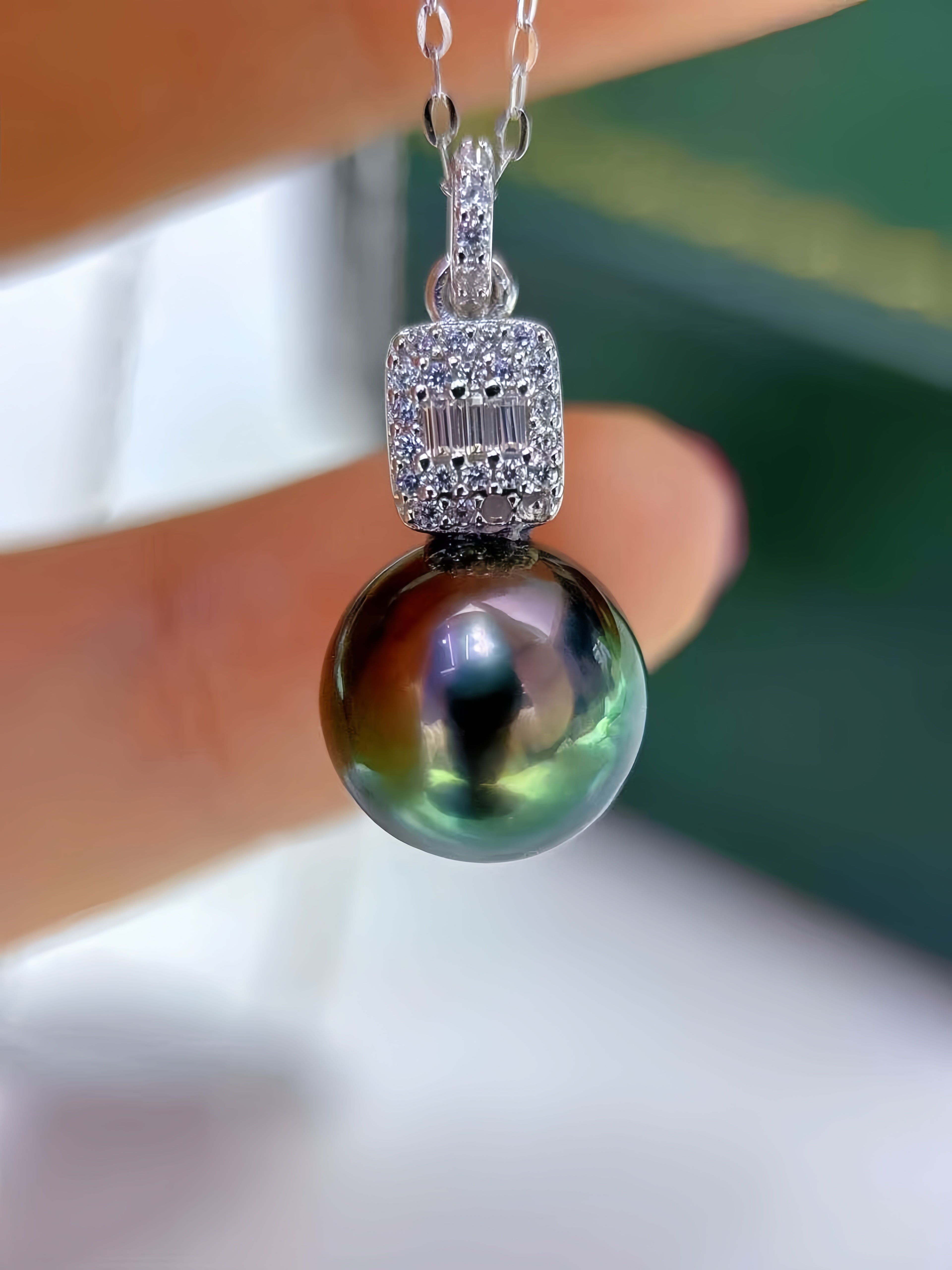 A close-up of a pendant necklace held by fingers, featuring a large iridescent dark pearl pendant with a silver-toned pavé bezel and chain.