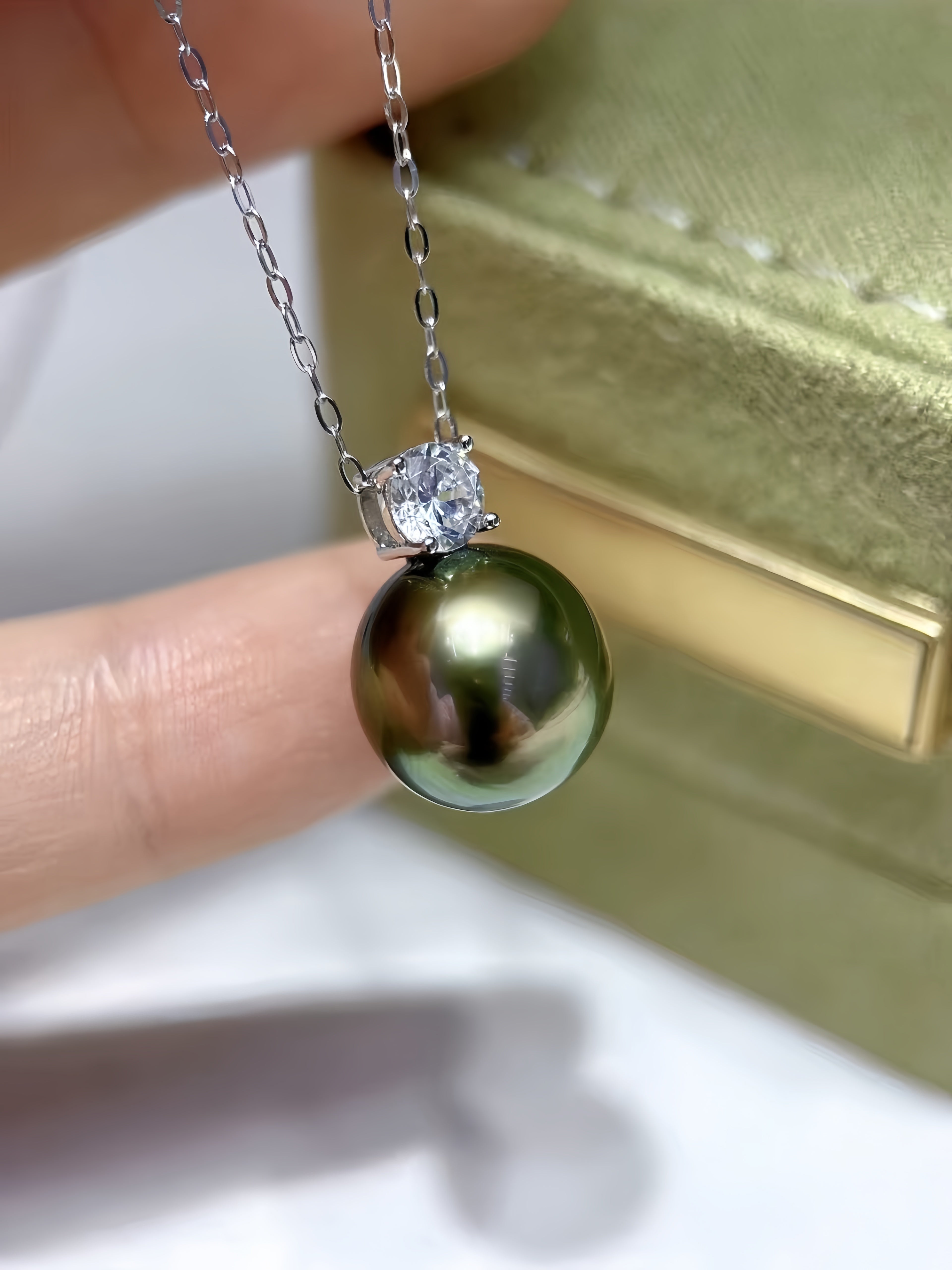 A close-up of a necklace with a round dark green pearl pendant and a clear stone setting on a silver chain, held between fingers with a gold jewelry box in the background.