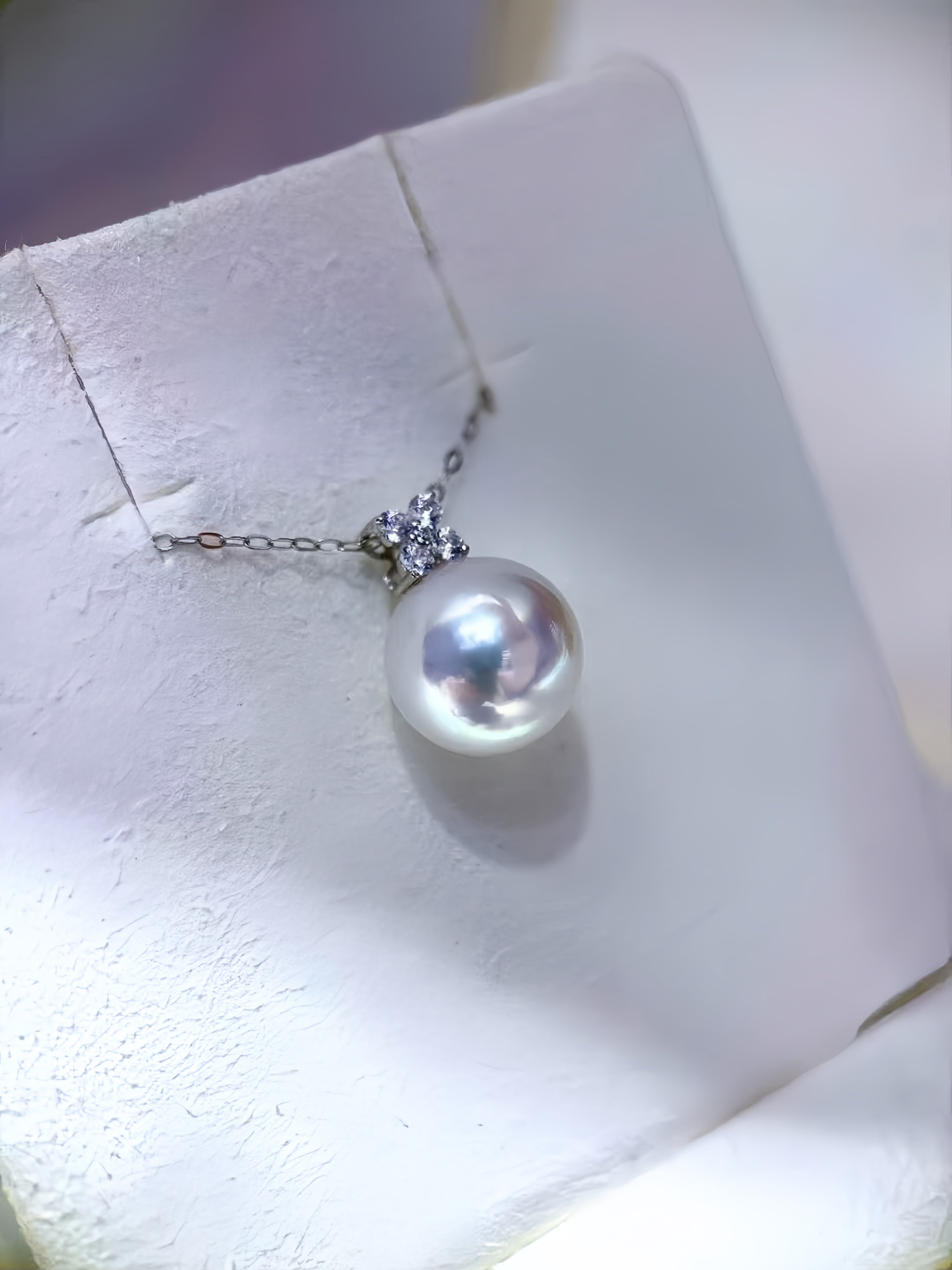 A single pearl pendant necklace with a small silver floral setting and chain, displayed on a white jewelry stand.
