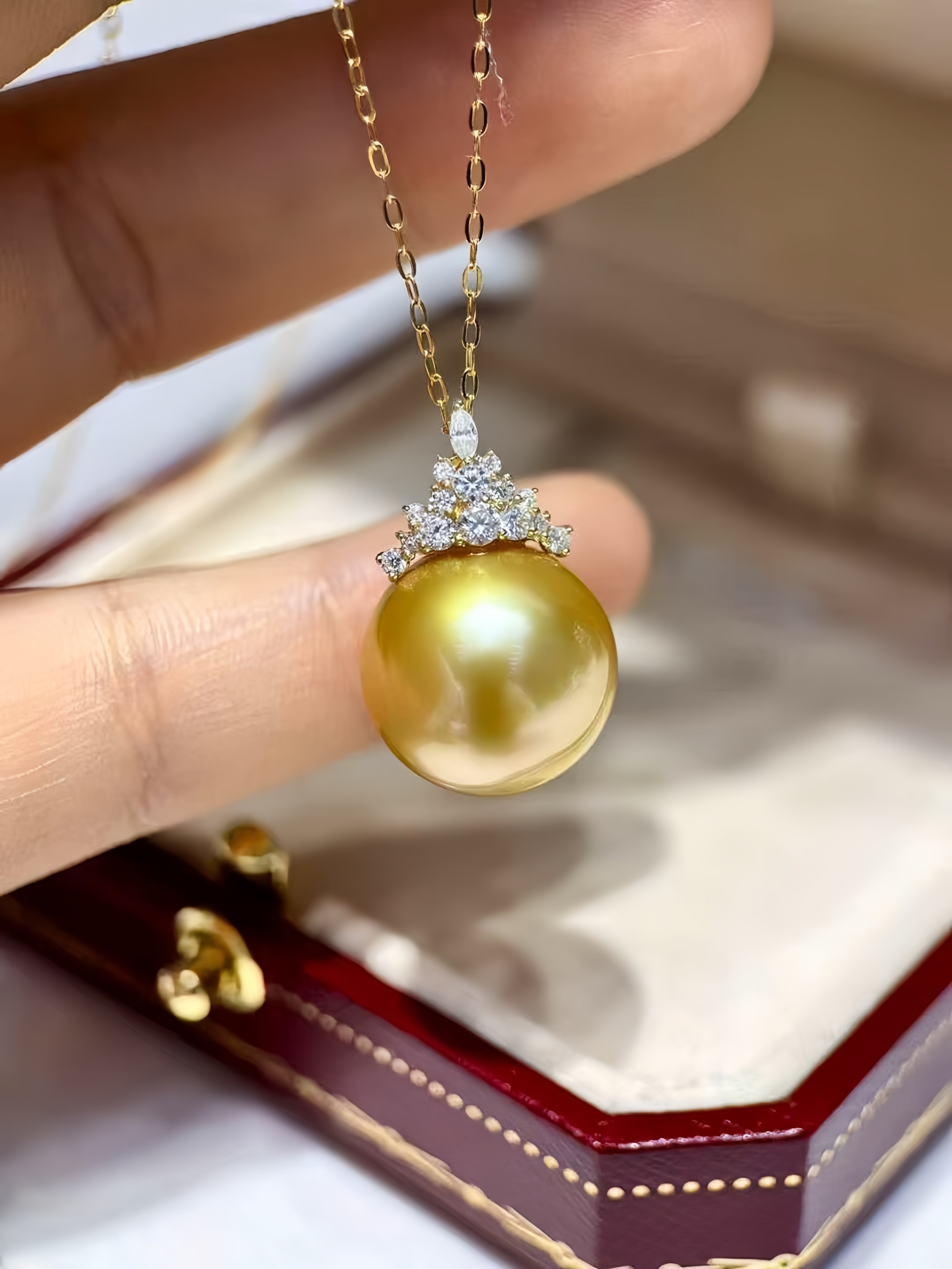 A close-up of a gold pendant necklace with a large round yellow-gold pearl and a small diamond crown setting, held by a hand above an open jewelry box.