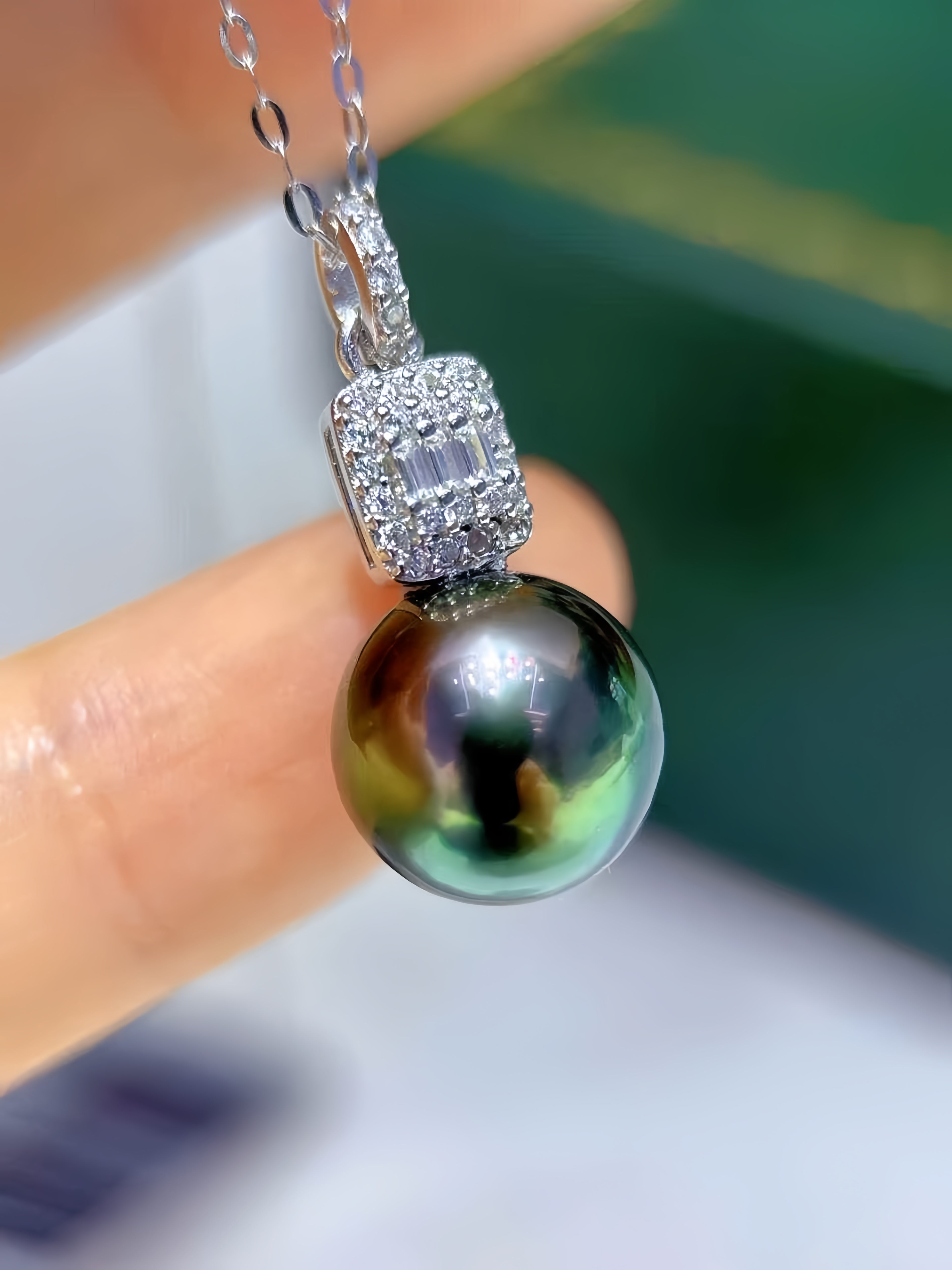A close-up of a pendant necklace held between fingers, featuring a large iridescent dark pearl suspended from a silver-toned chain with a pavé-set rectangular bezel.