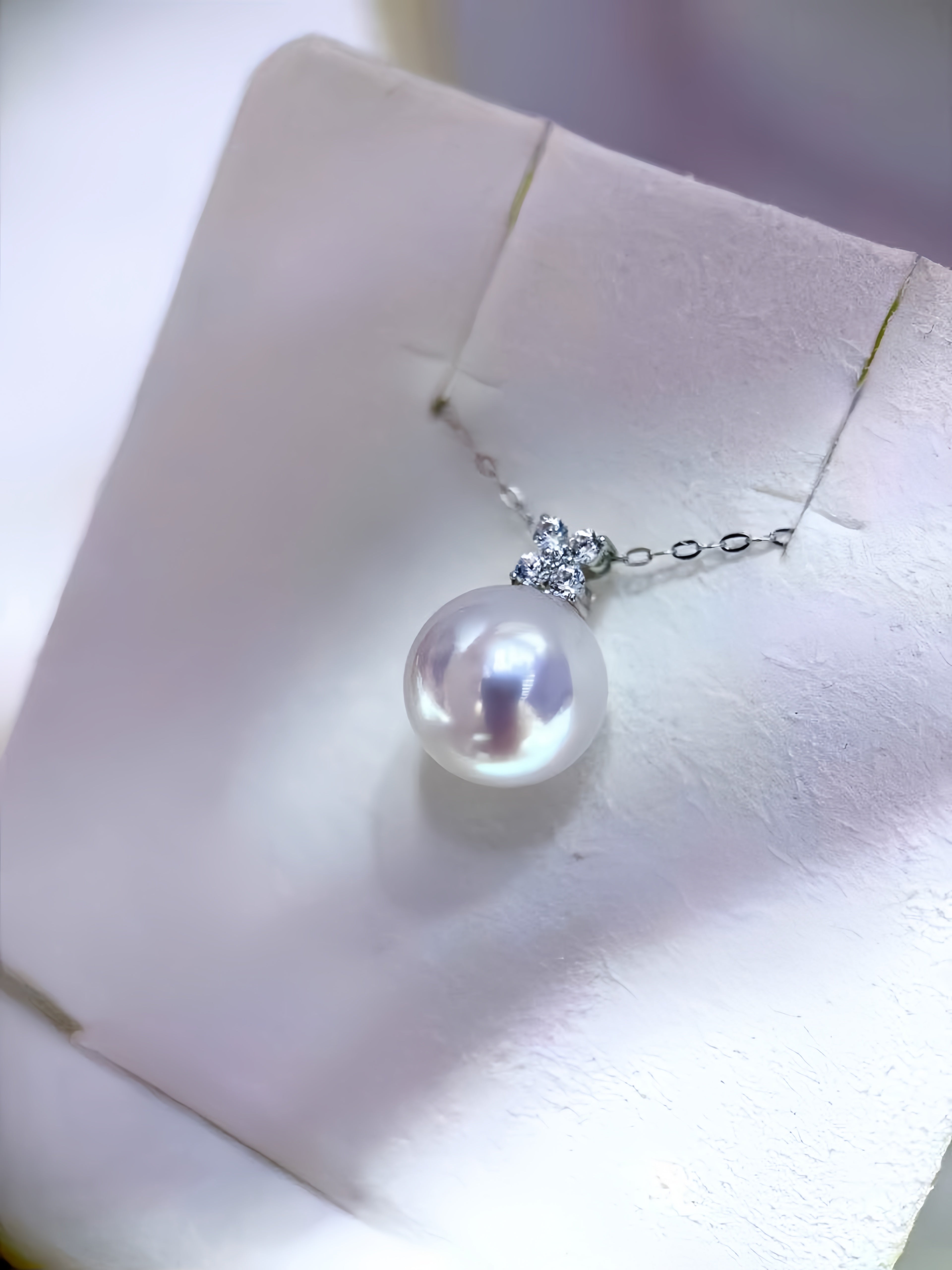 A close-up of a pearl pendant necklace with a silver-toned chain and a small clear stone cluster, displayed on a white jewelry stand.